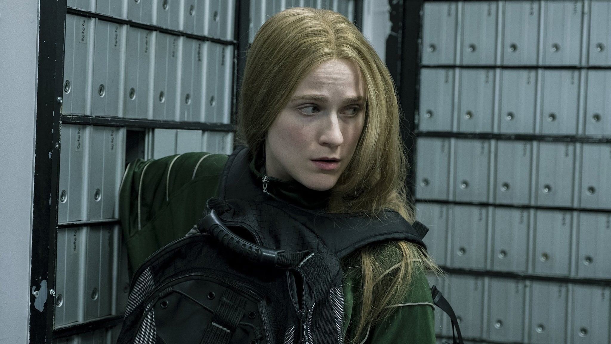 A person with long blonde hair, wearing a backwards backpack, gazes away and looking worried, against a backdrop of metal lockers.