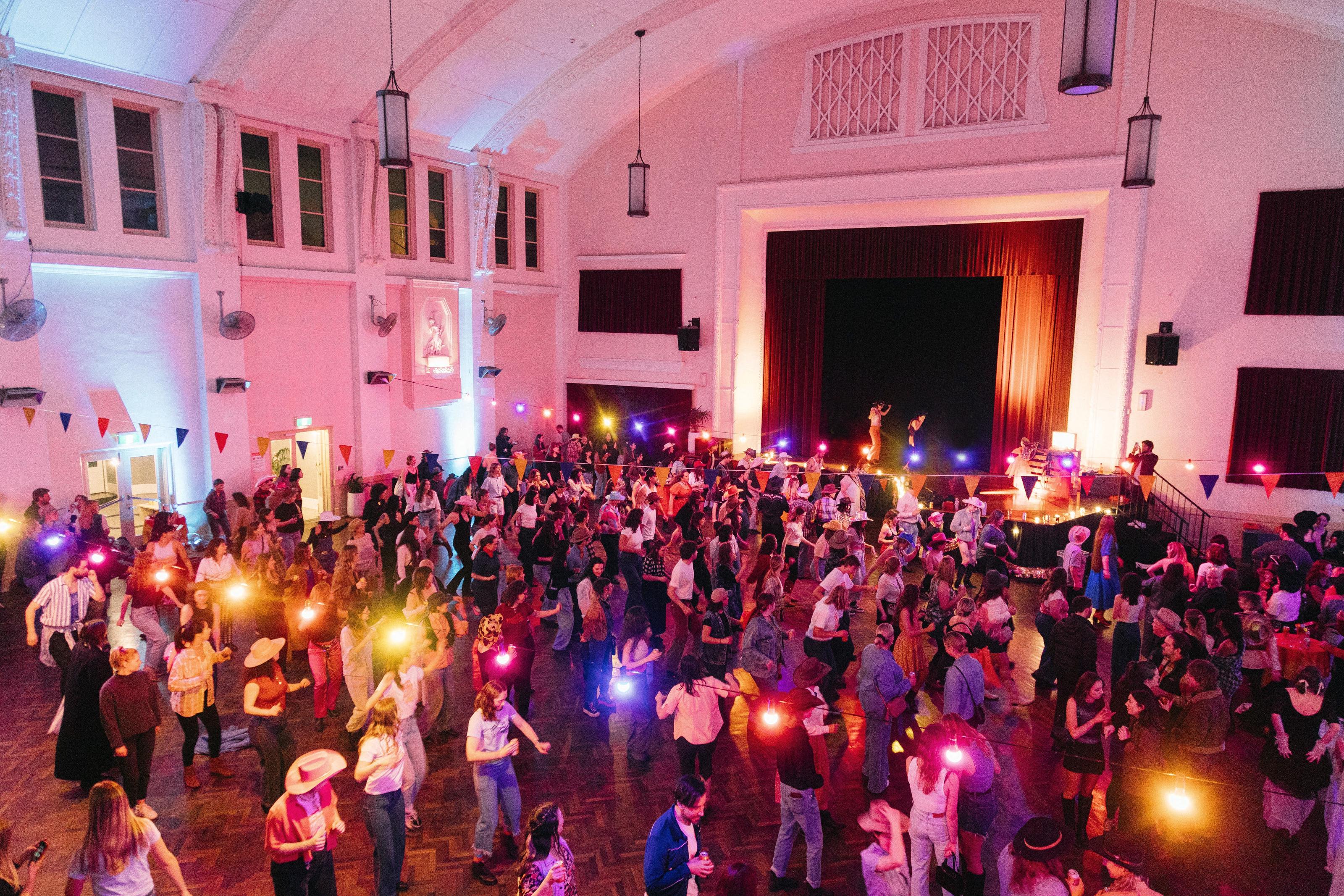A large crowd gathered together, dancing in a colourfully lit hall.