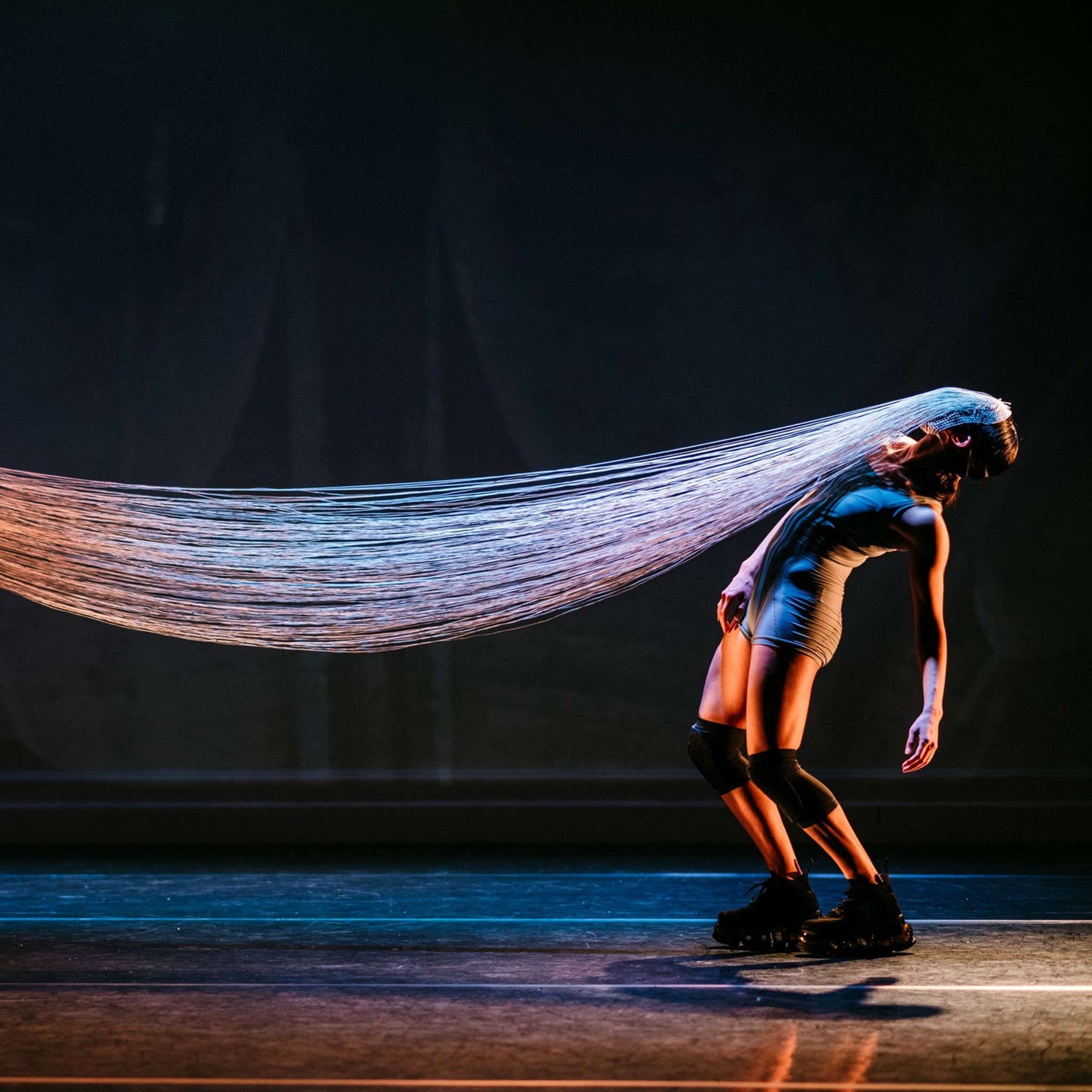 A dancer in knee pads arching backward, her face attached to a long, flowing strands of fabric.