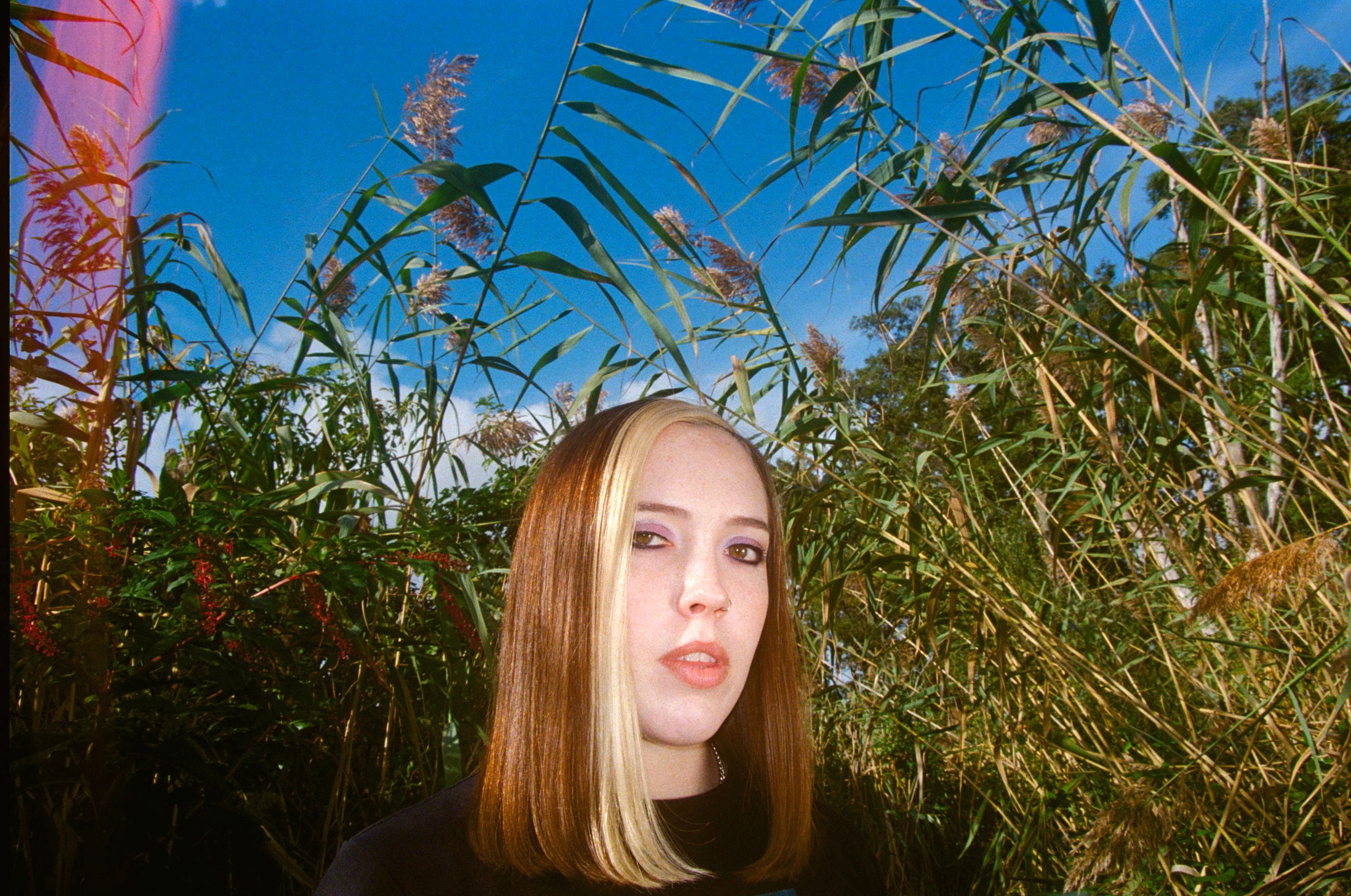 A mid shot of person with shoulder length hair looking into the camera. Behind them are flowering bushes. 