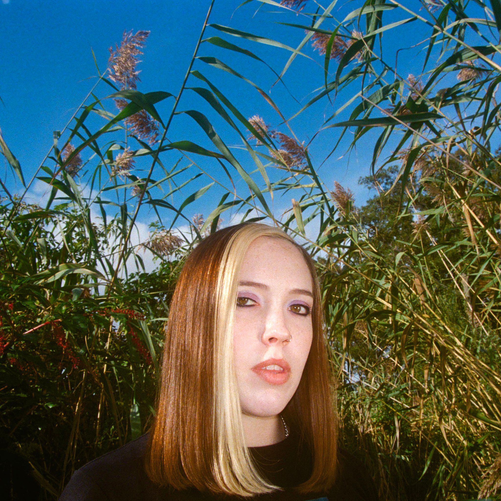 A mid shot of person with shoulder length hair looking into the camera. Behind them are flowering bushes.