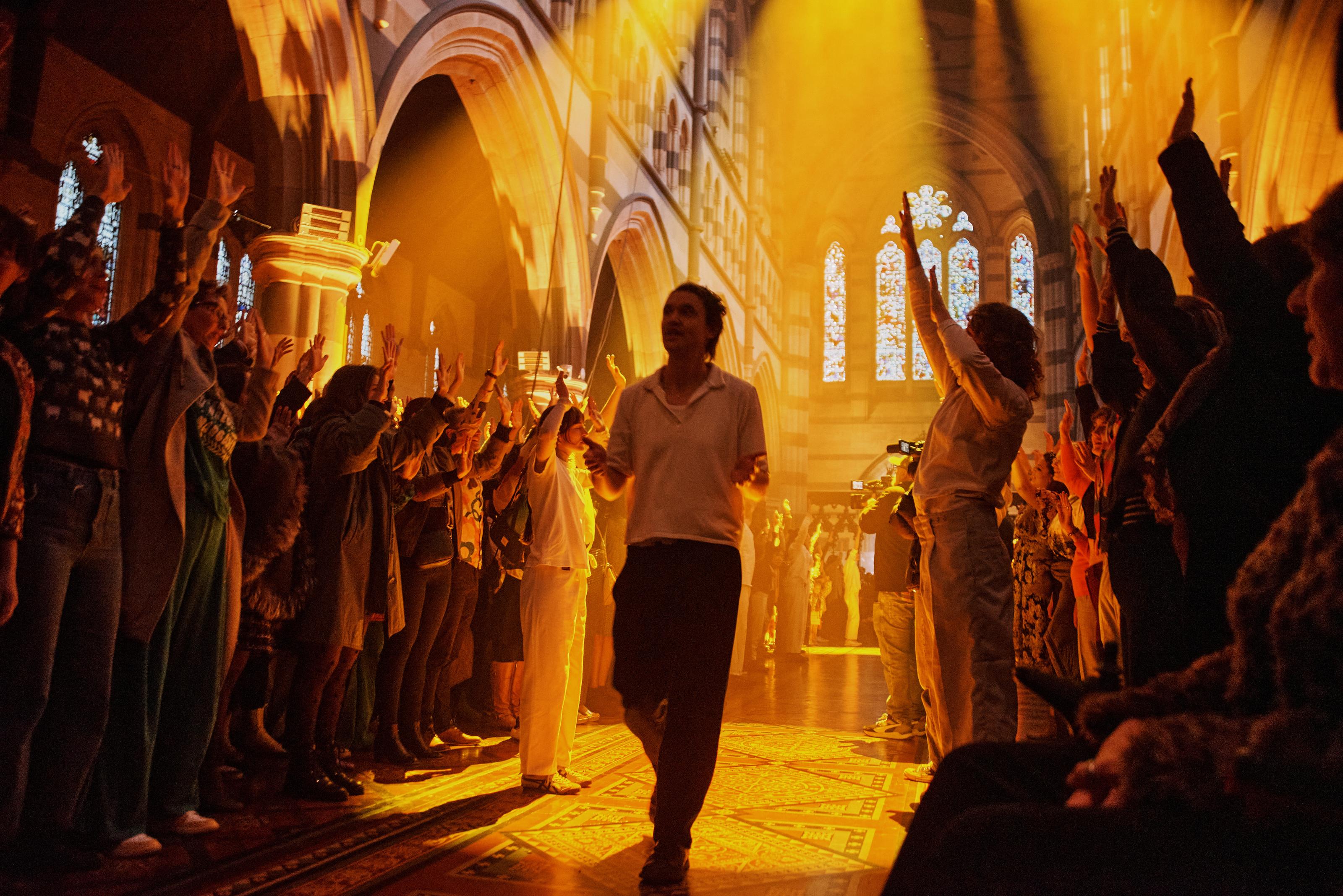 Two groups of people raise their arms in the air in unison, as a man walks between them singing