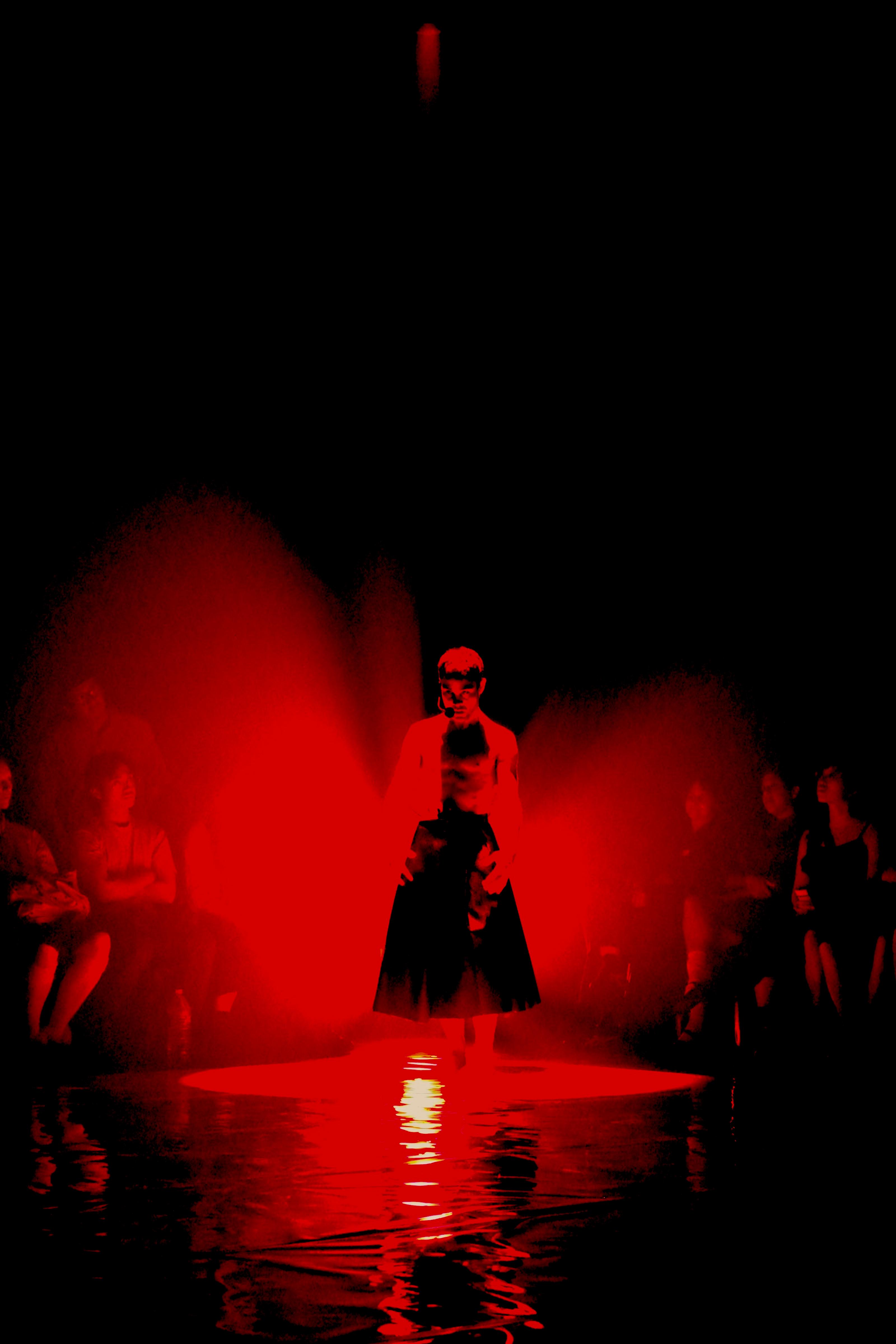 A performer wearing a long skirt standing in the middle of a light on stage–the image has been colour graded to increase the contrast of light and dark
