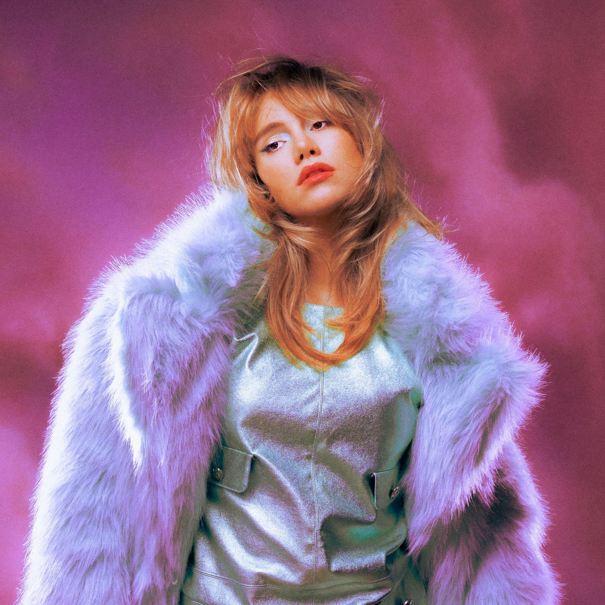 A portrait of Suki Waterhouse looking away from the camera, with soft purple clouds in the background.