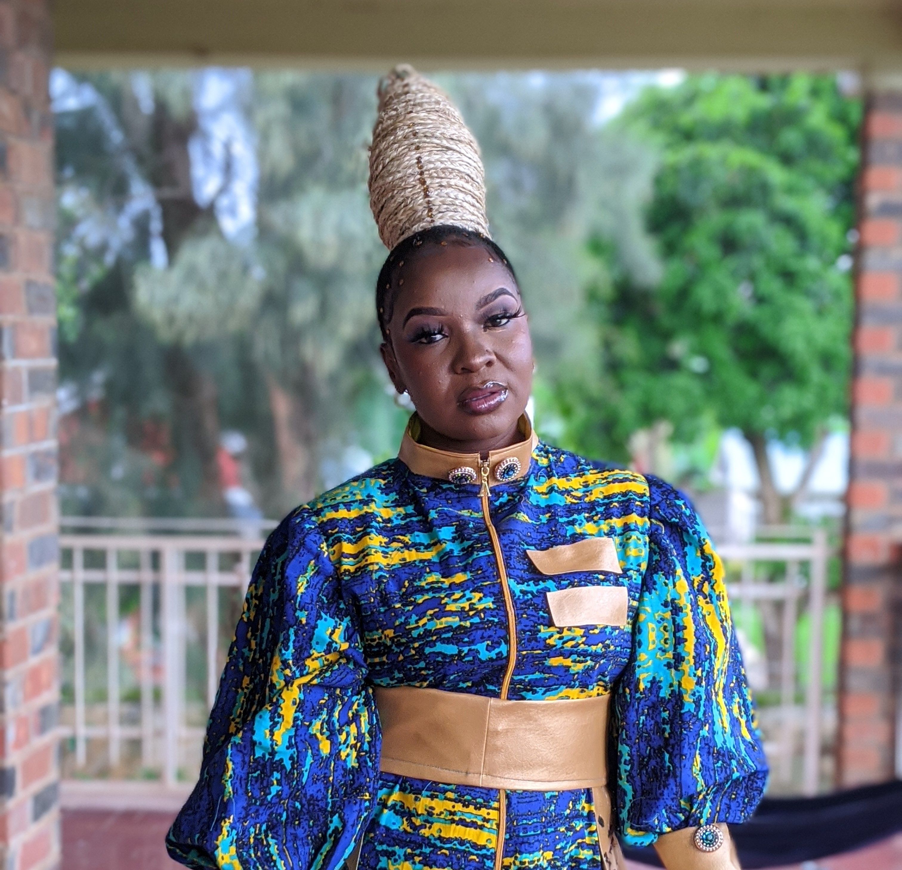 A person stands on a patio wearing a colorful patterned outfit with gold accents and a tall braided hairstyle.