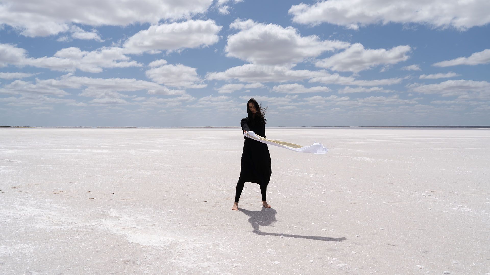 Positioned in a white and barren landscape, a woman dressed in black, with white fabric wrapped around her hand, stands agaze in the wind. 