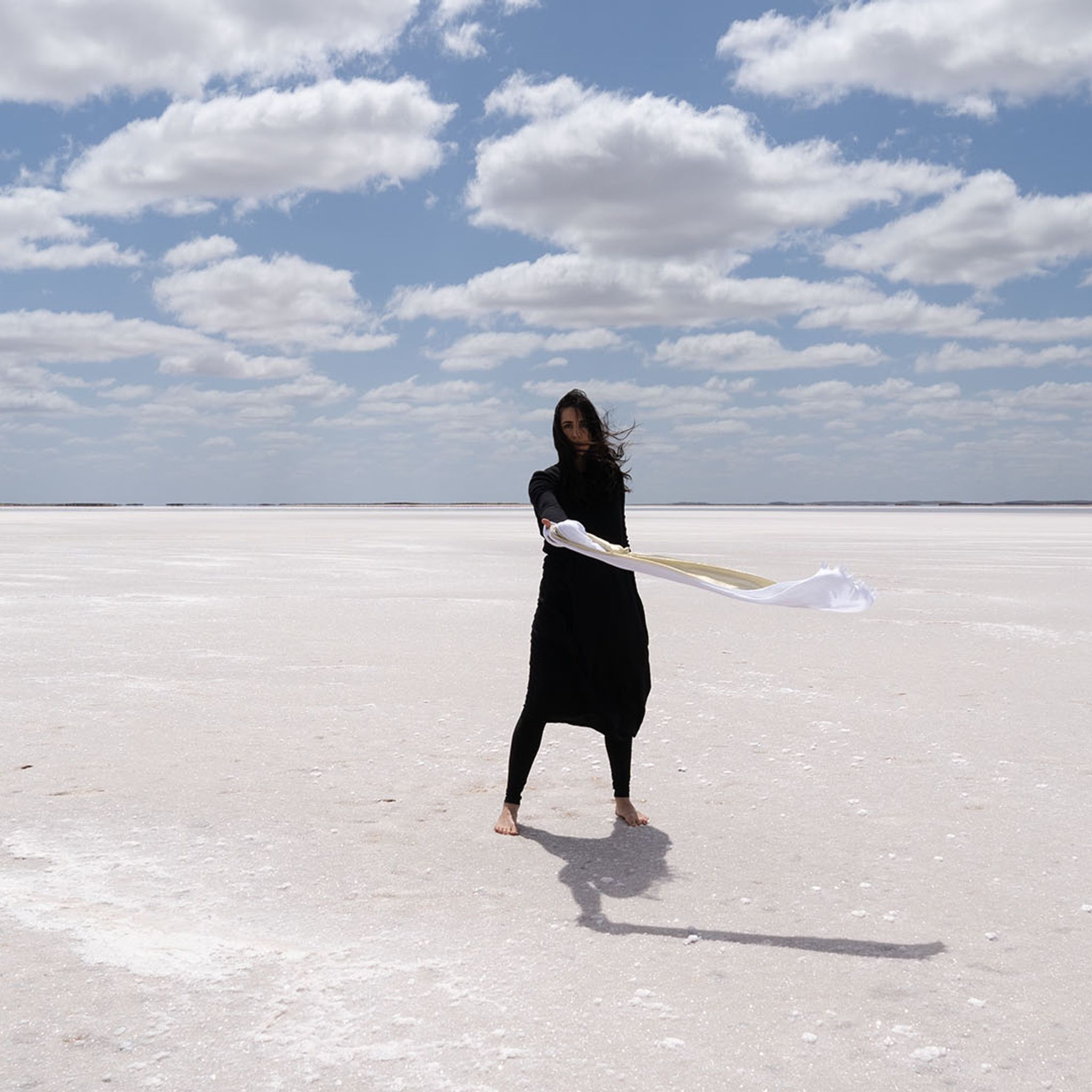 Positioned in a white and barren landscape, a woman dressed in black, with white fabric wrapped around her hand, stands agaze in the wind.