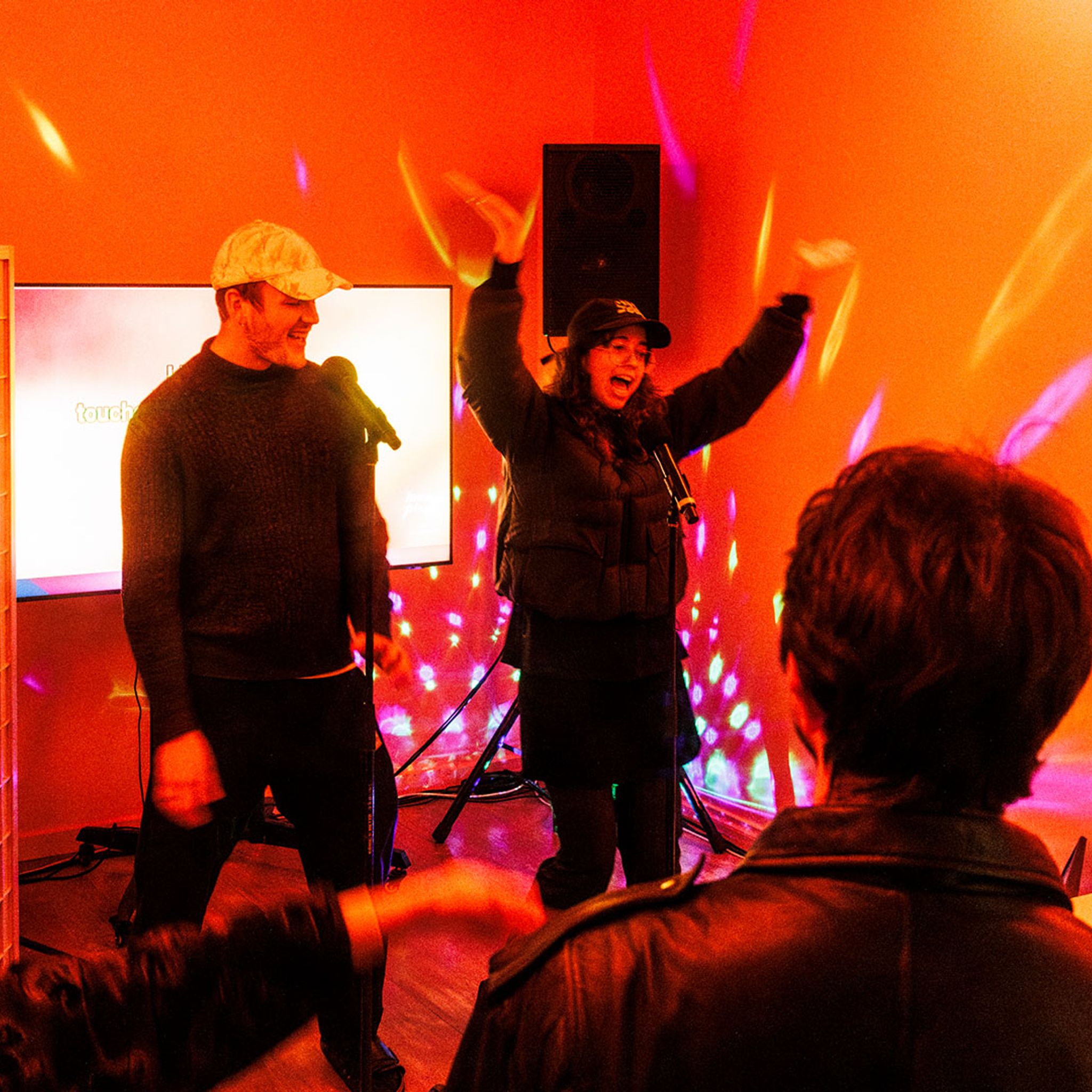 Two people perform enthusiastically with a microphone in a vibrant, orange-lit room as a small crowd watches and records.