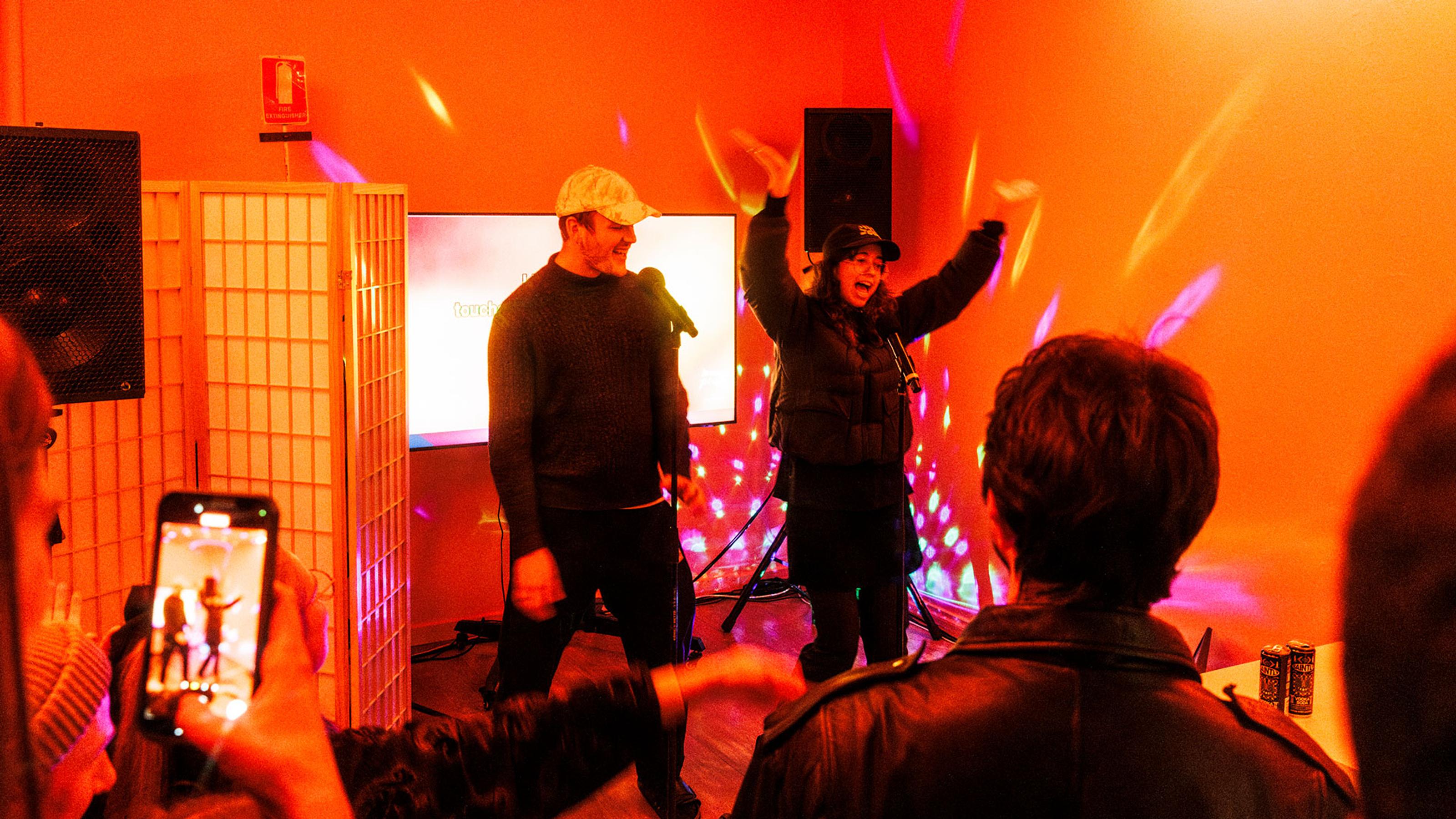 Two people perform karaoke for people in a room lit up with disco lights.