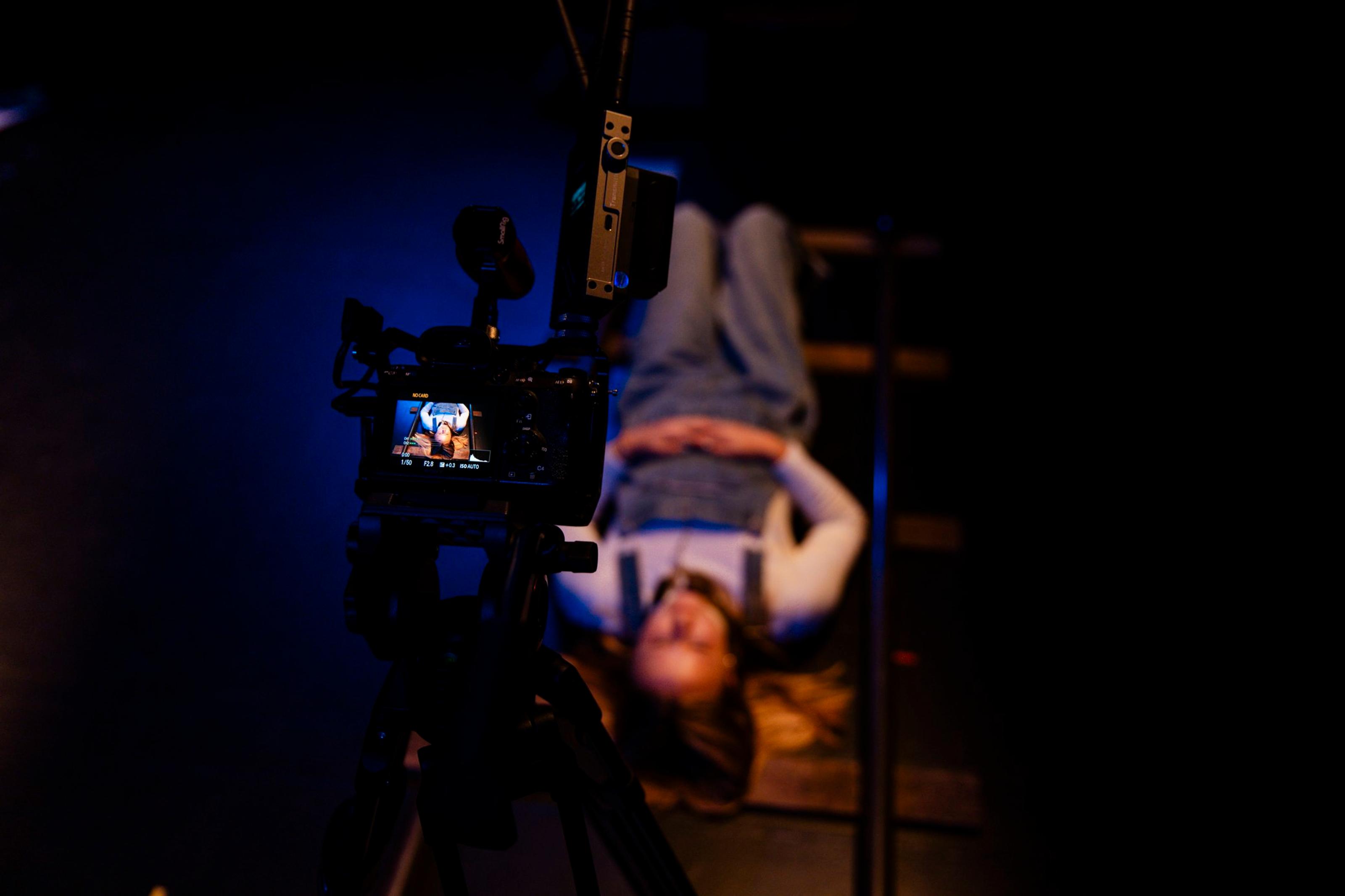 A young person hangs upside down like they’re on monkey bars. They are filming themselves with a camera