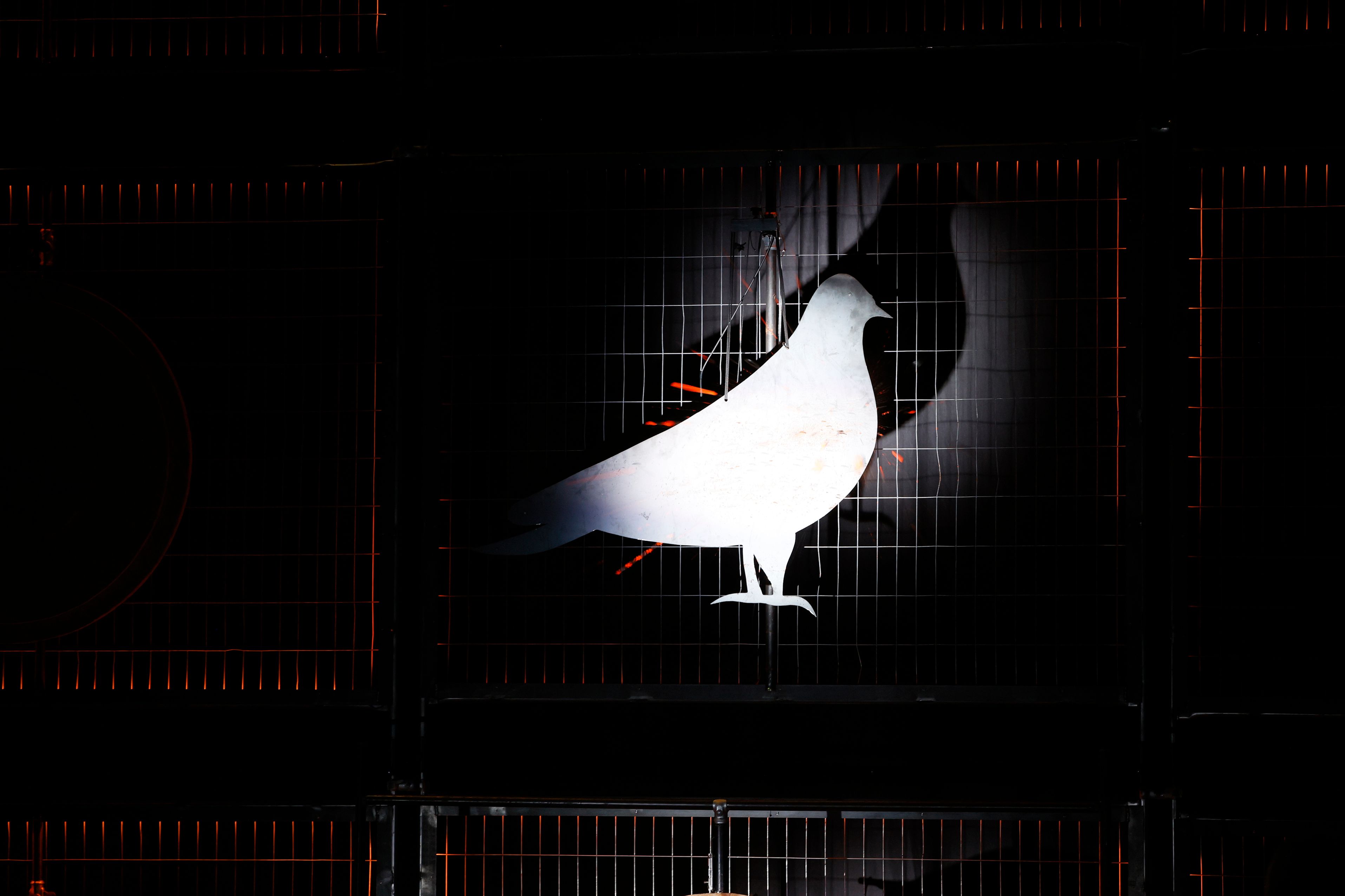 In a dark space, a spotlight reveals a metal silhouette of a pigeon, hanging from a wired cage. 
