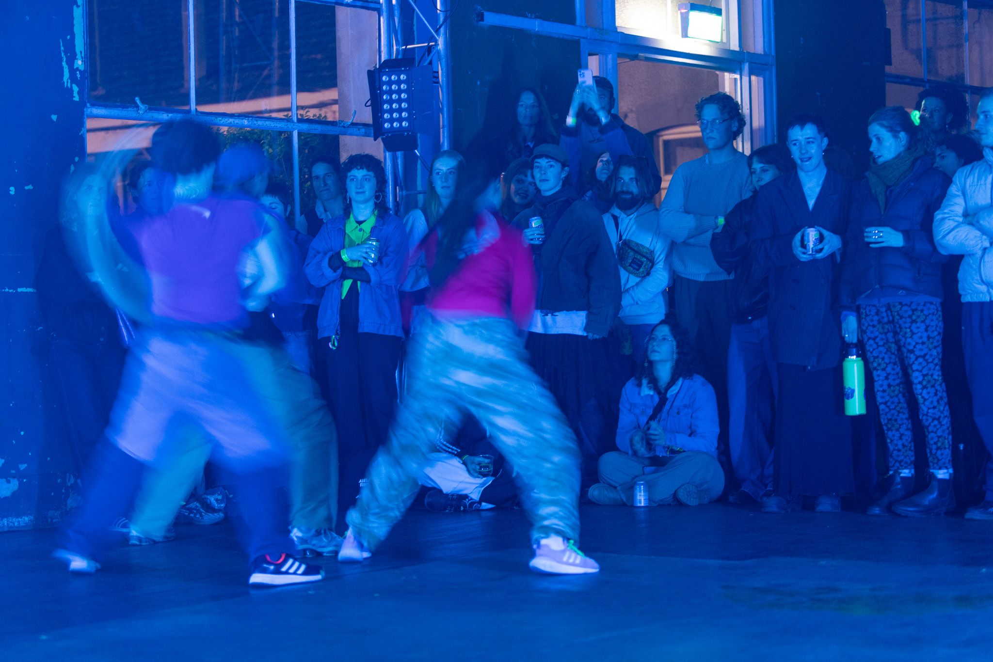 A trio of dancers perform close to a standing and sitting crowd, with their motion blurred. 