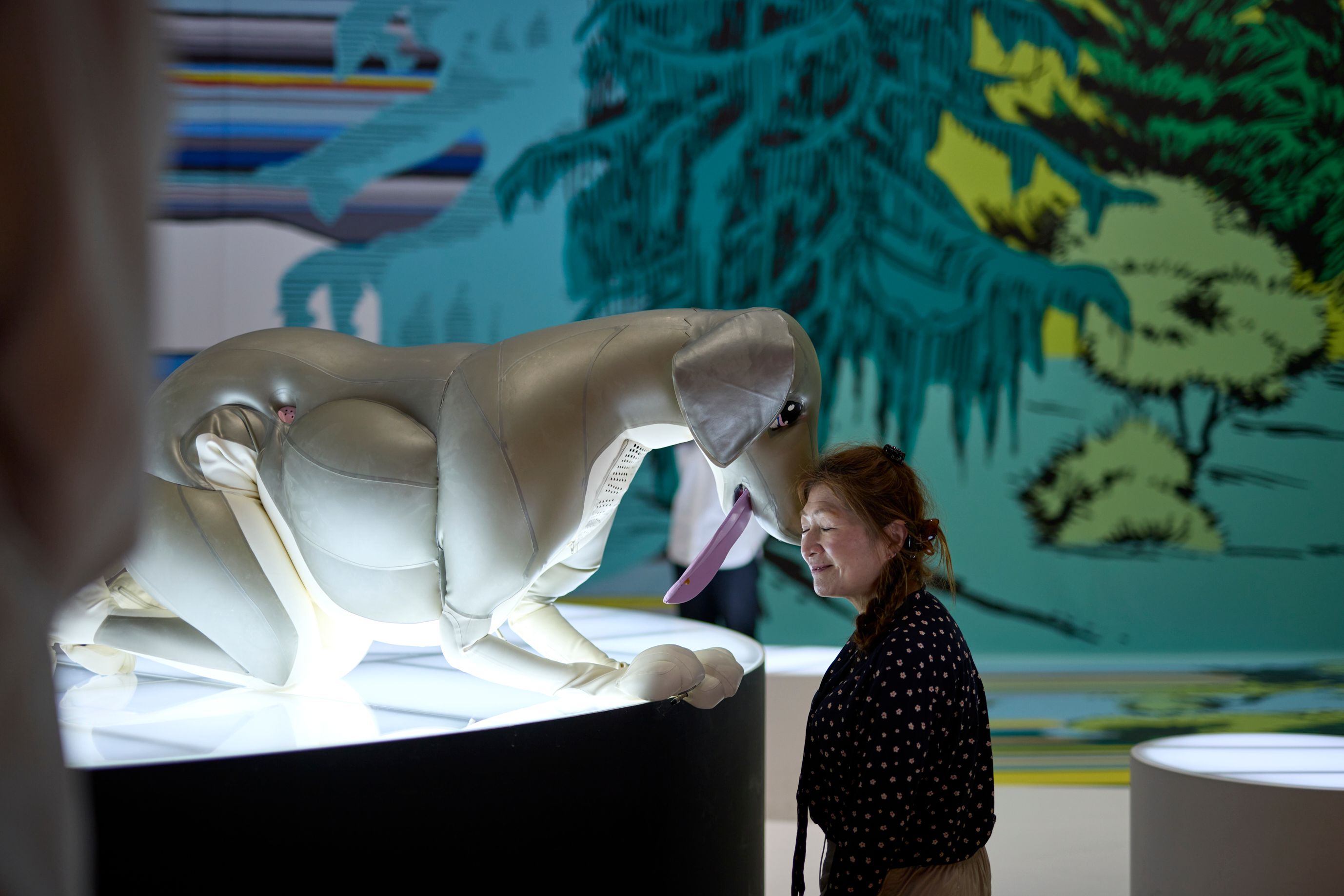 A woman lovingly places her head against the snout of a large inflatable dog, which is crouched on all fours, sticking its tongue out.  