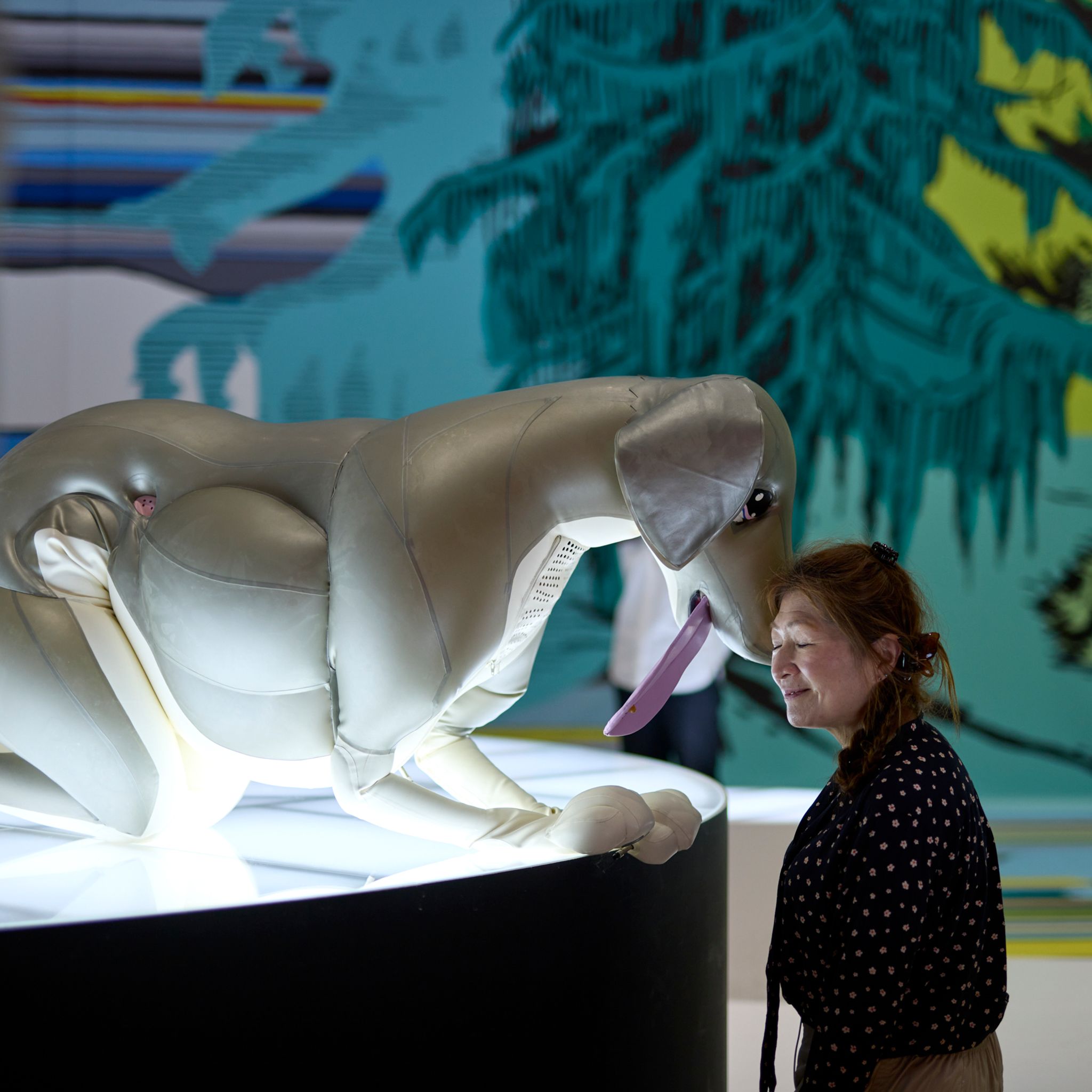 A woman lovingly places her head against the snout of a large inflatable dog, which is crouched on all fours, sticking its tongue out.