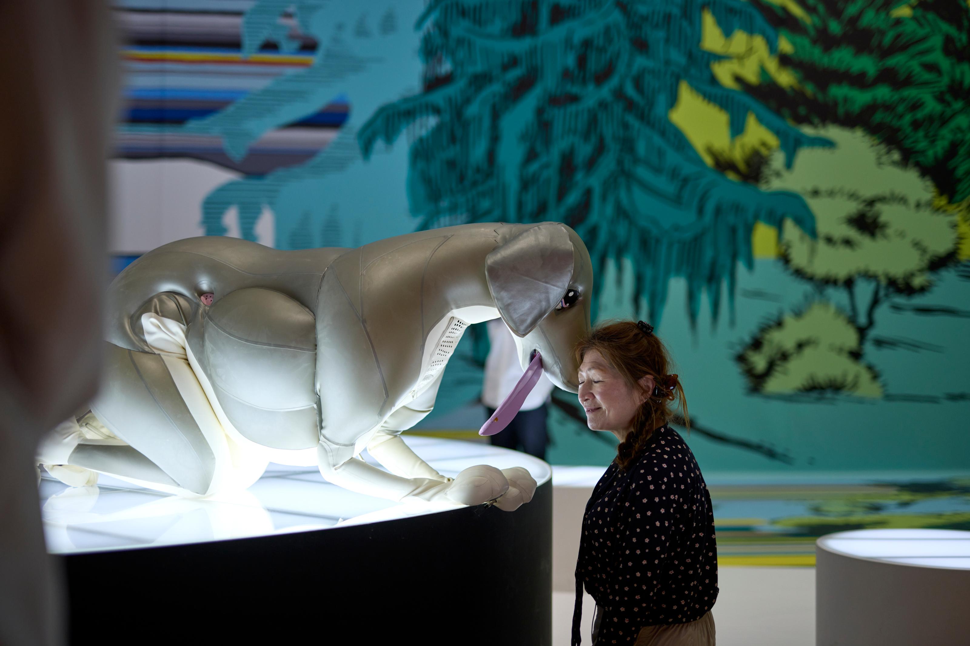 A woman lovingly places her head against the snout of a large inflatable dog, which is crouched on all fours, sticking its tongue out.