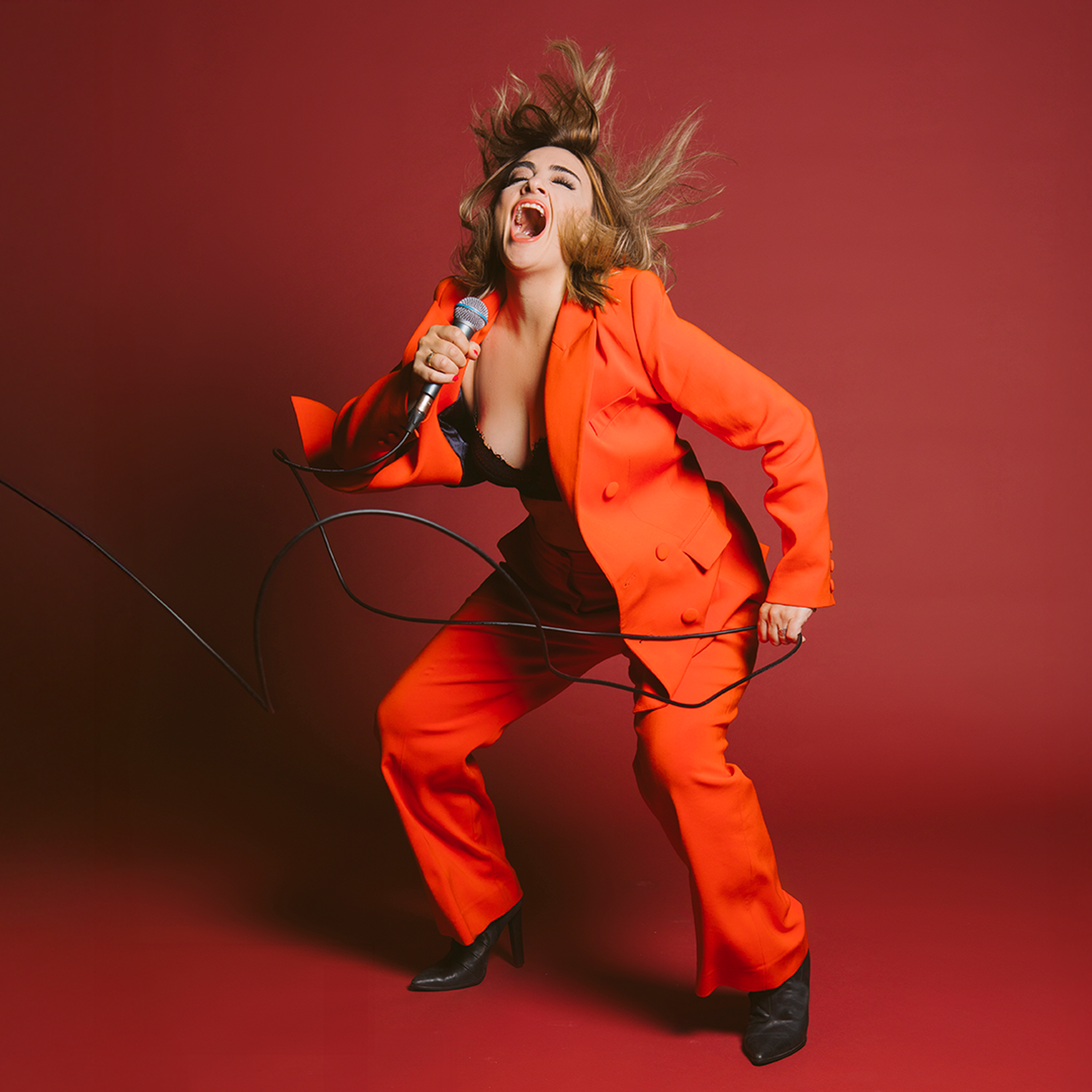 A woman in an orange suit, mid-singing performance, holding a mic and hair flying