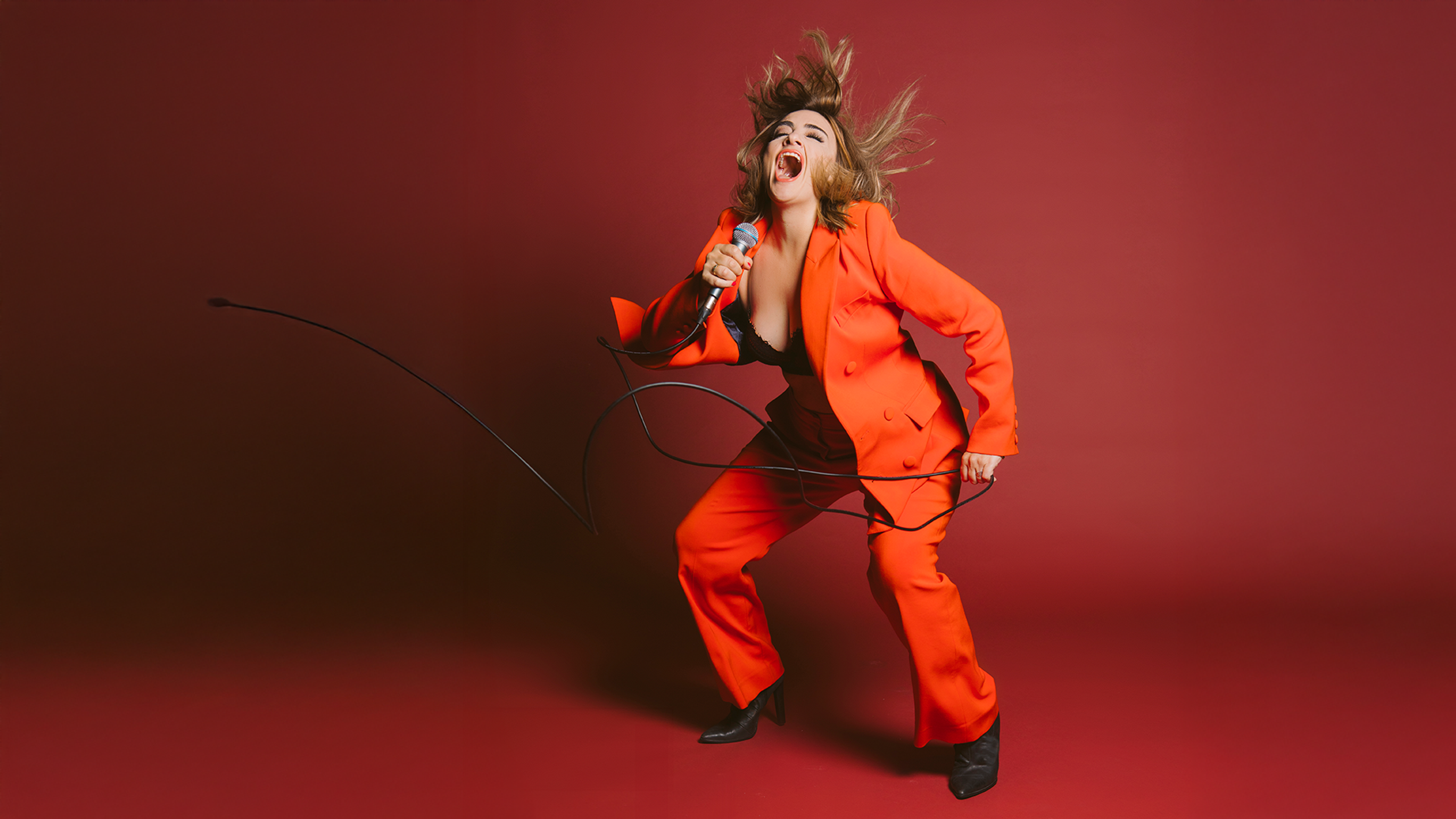 A woman in an orange suit, mid-singing performance, holding a mic and hair flying