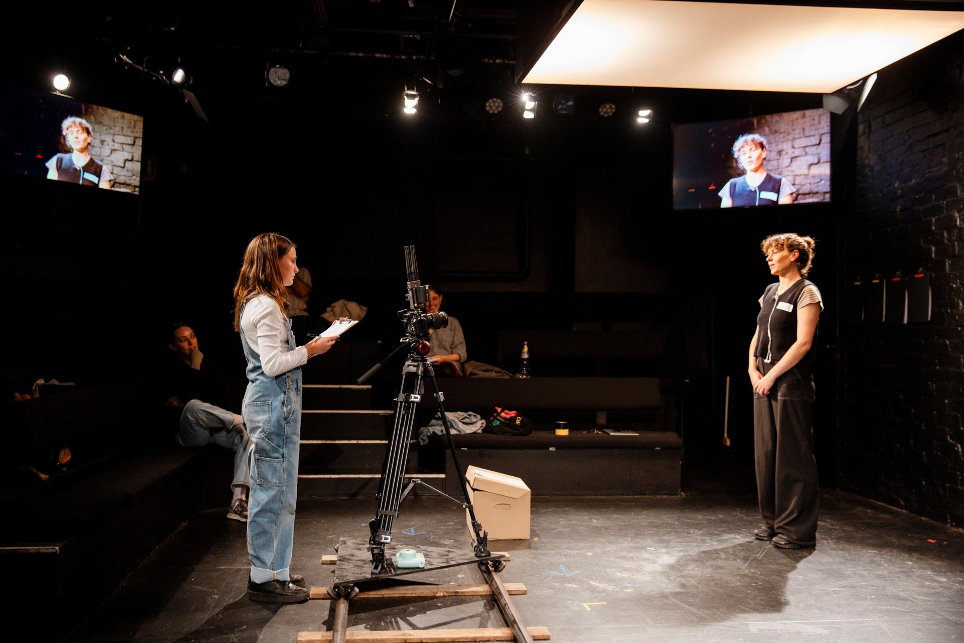 A young person holding a script and standing next to video camera films an adult person, the video is being livestreamed to a screen