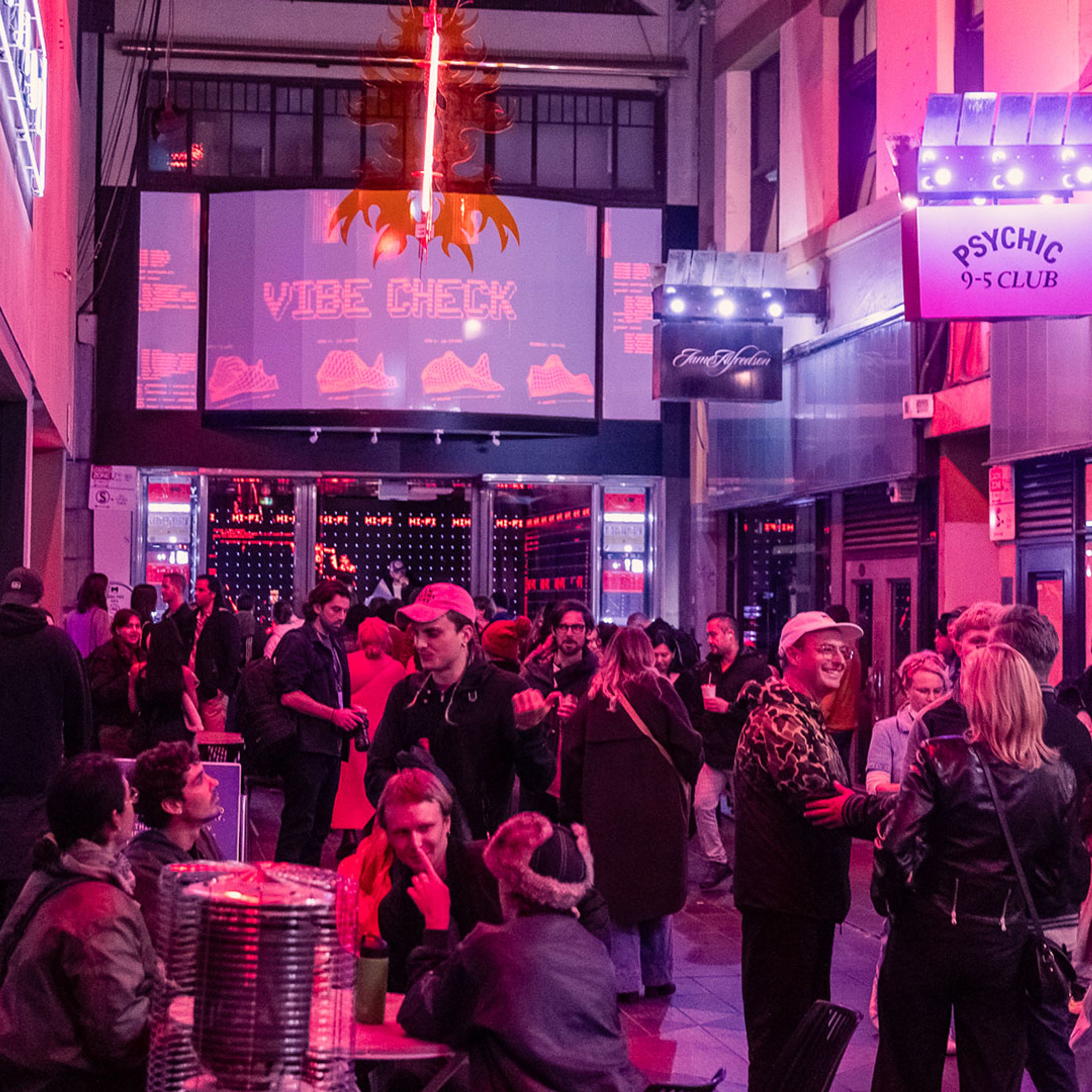 A wide view of a busy arcade at night. People are enjoying drinks and listening to a DJ