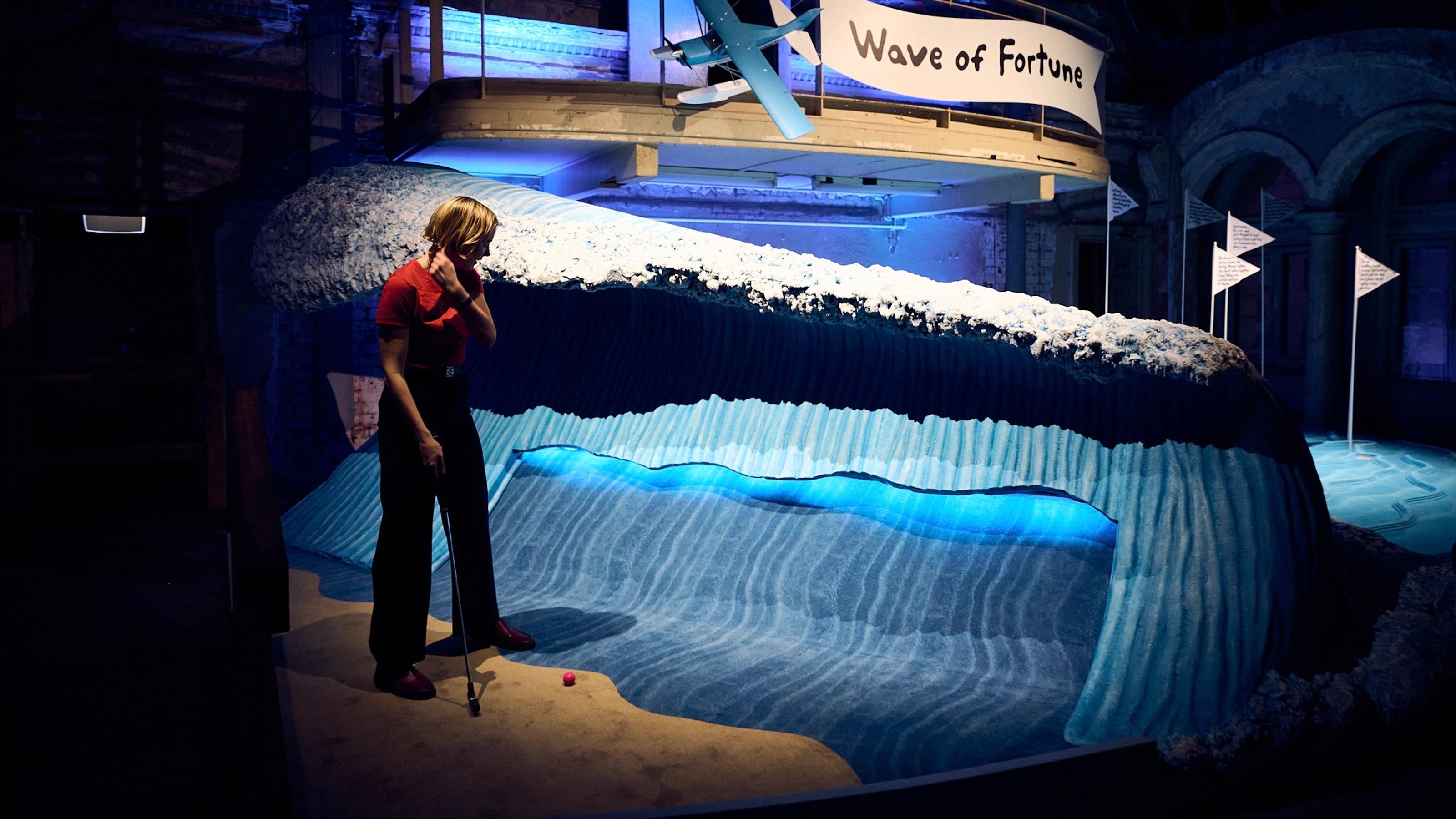Someone about to try Miranda July's Wave of Fortune mini-golf hole, the obstacle is a big foamy wave.