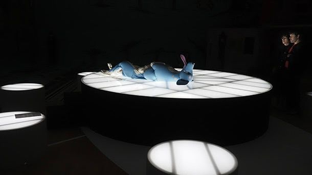 Four white elevated round tables light up a dark room. On the centre table, a large inflatable blue dog lies on its back.