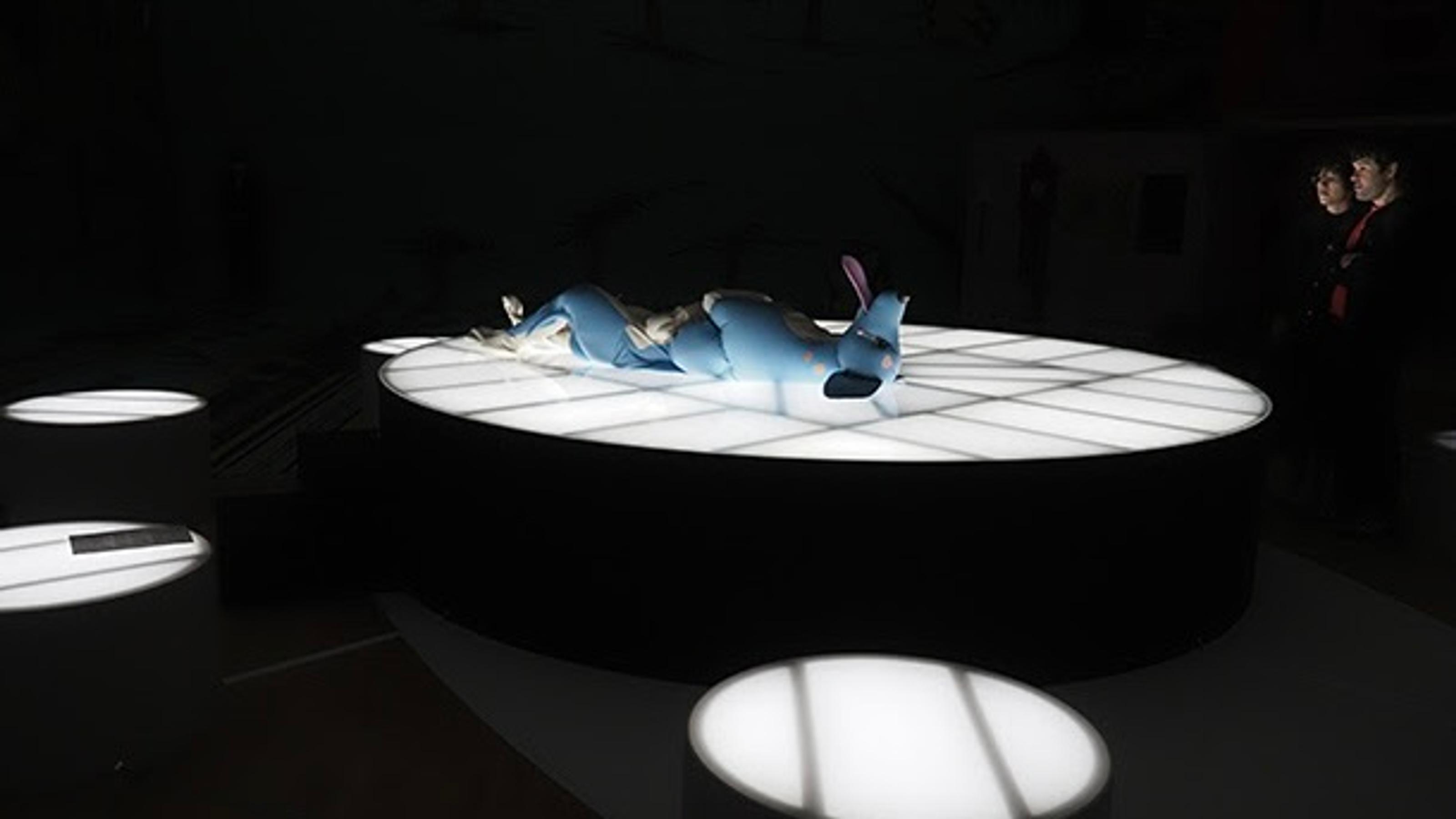 Four white elevated round tables light up a dark room. On the centre table, a large inflatable blue dog lies on its back.