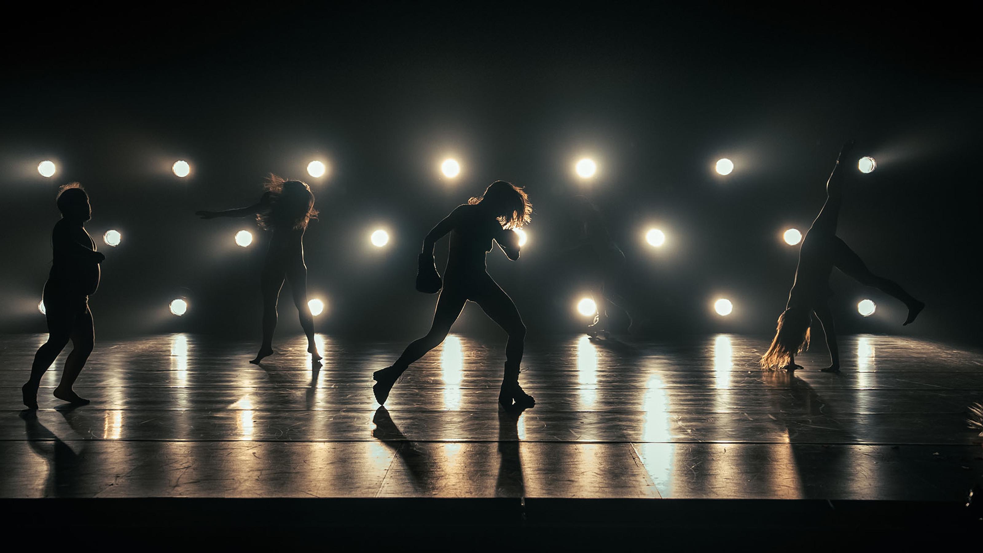 Four nude performers are backlit and silhouetted by a bank of search lights