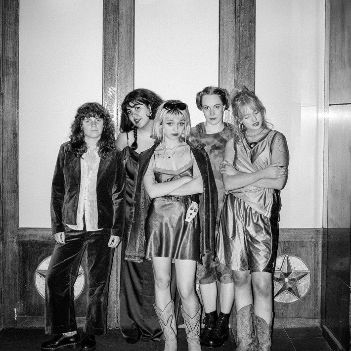 Five members of The Vovo's in black and white: two at the front who wear short dress, crossing their arms—one in the center with sunglasses on her head and the other with her eyes closed. The one on the far-left wears trousers, the one on the far-right wears shorts, and the one in the back wear long dresses.