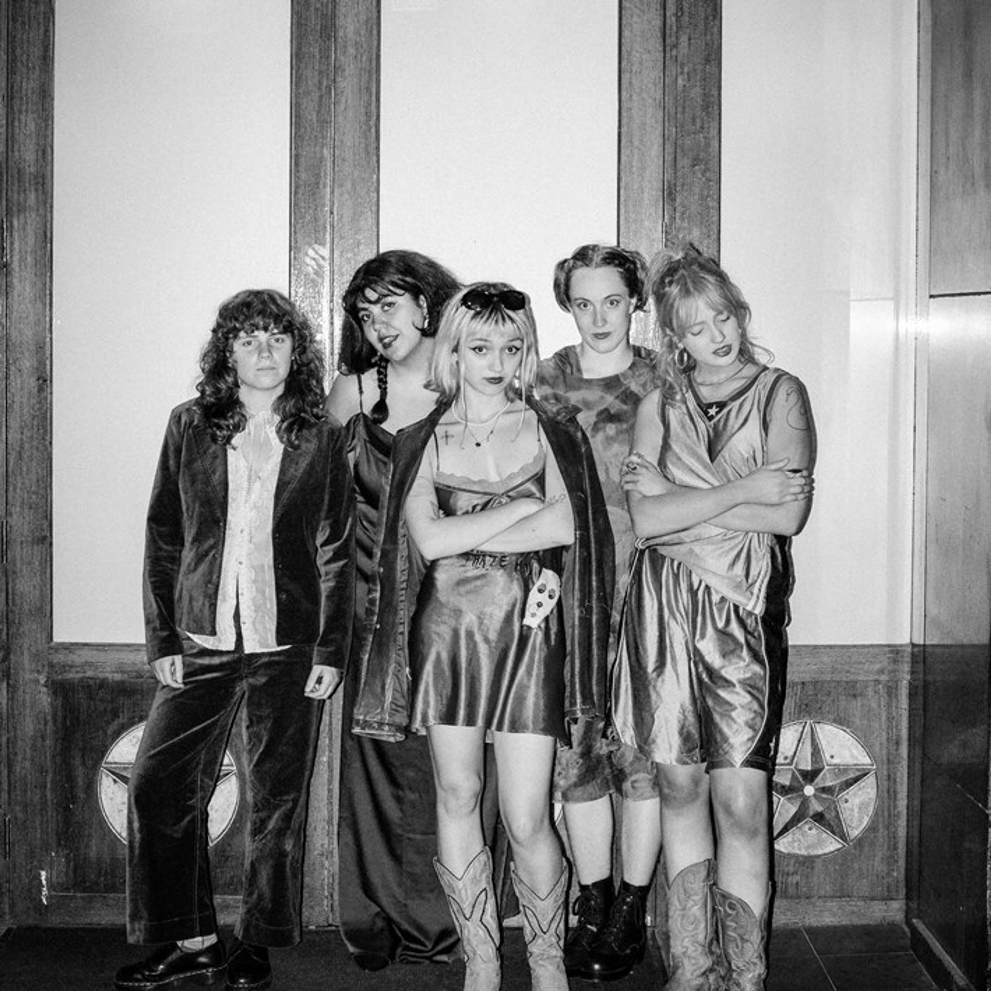 Five members of The Vovo's in black and white: two at the front who wear short dress, crossing their arms—one in the center with sunglasses on her head and the other with her eyes closed. The one on the far-left wears trousers, the one on the far-right wears shorts, and the one in the back wear long dresses.