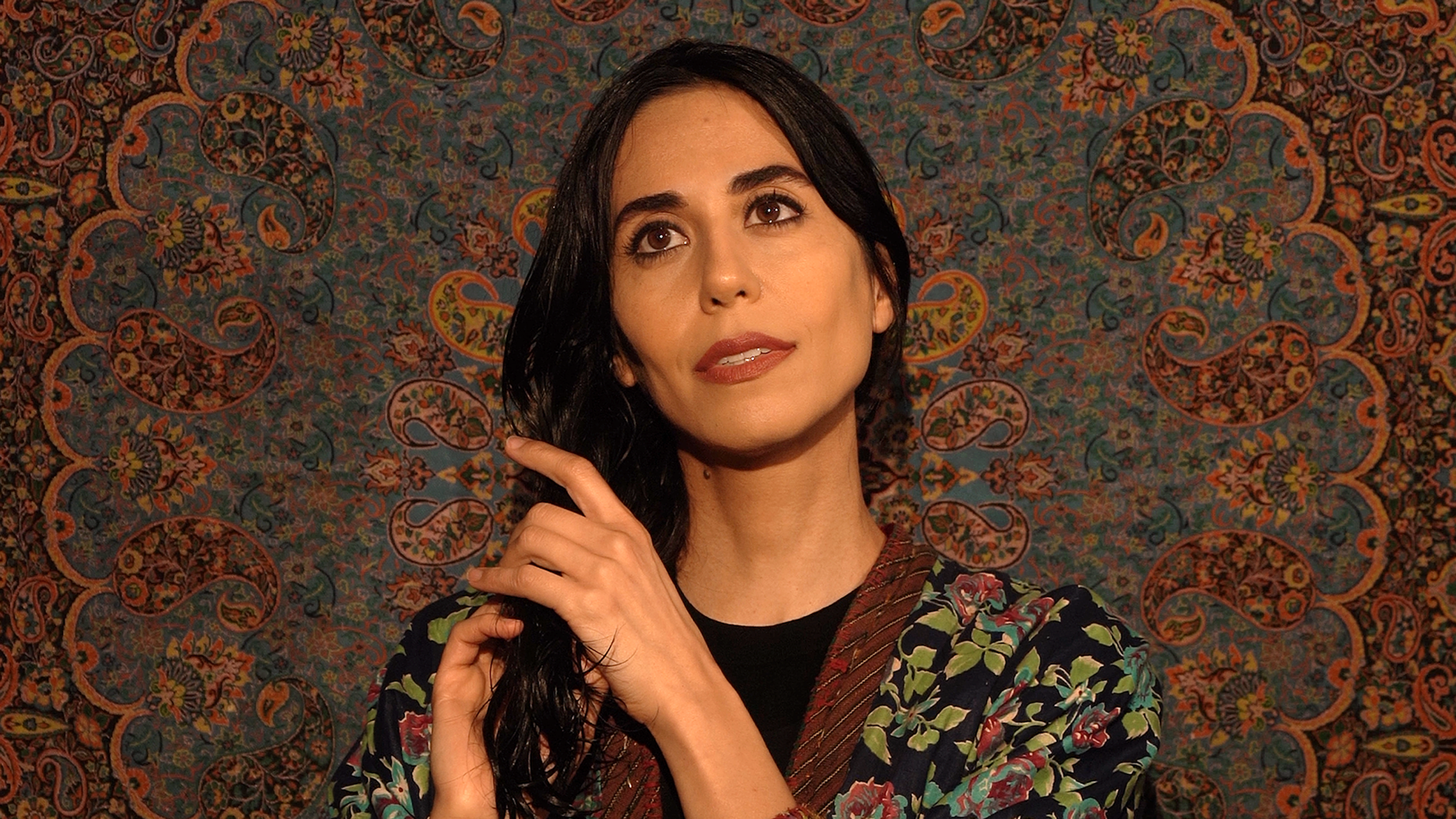 In front of a large patterned screen, a woman plays with her long, dark hair while gazing upwards.