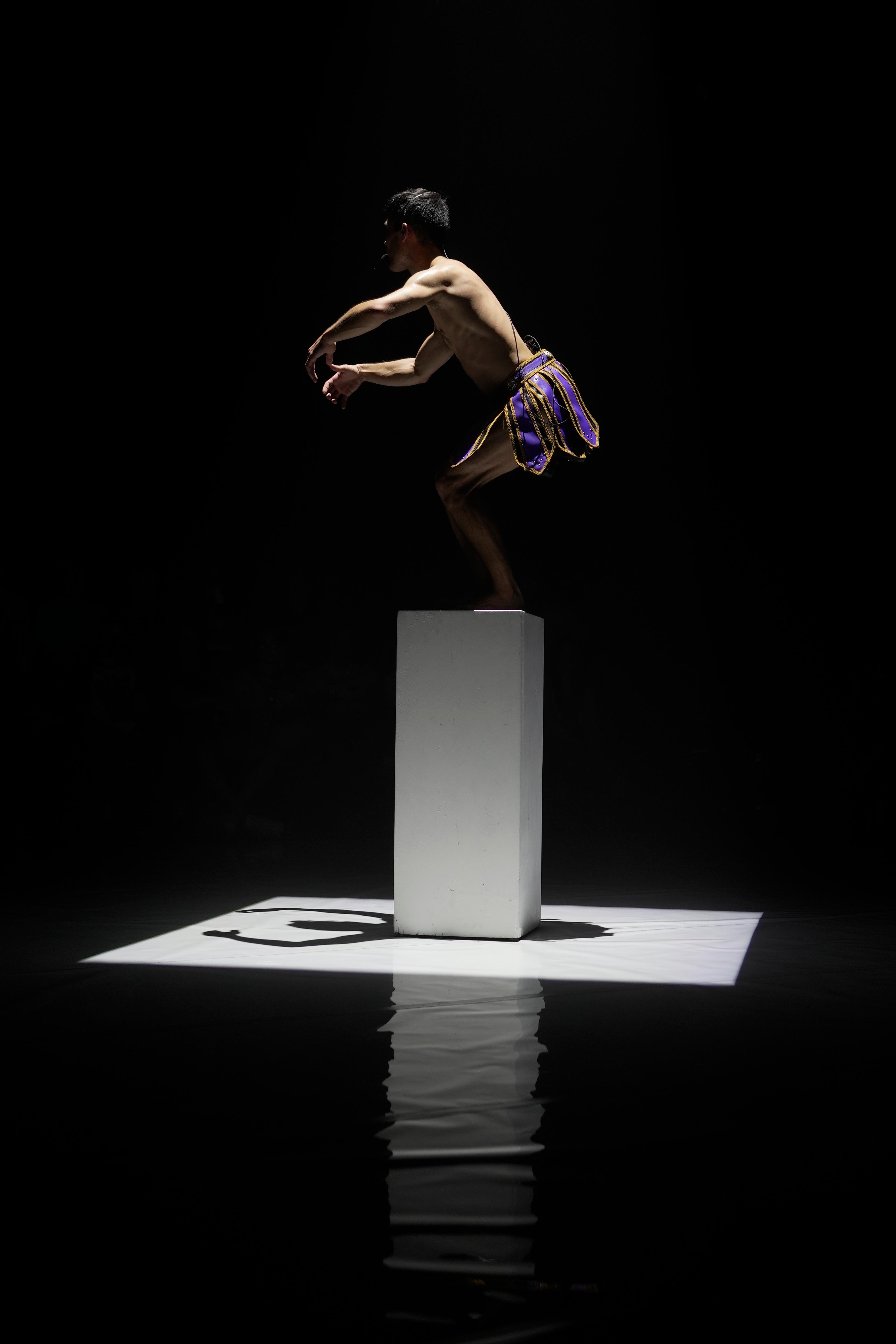 A performer in a skirt poses on top of the podium as a square spotlight is cast down upon them.
