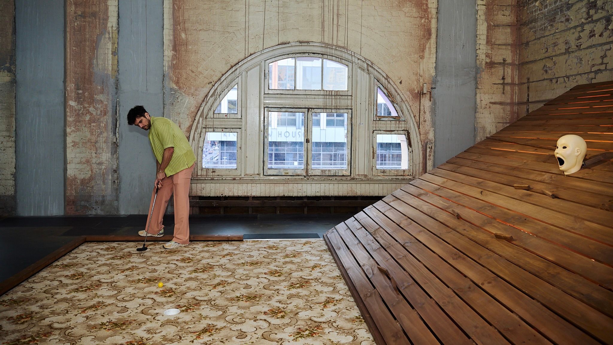 A sculpture of a head with a. wide-open mouth opens onto a sloping wooden deck. A person lines up to hit the ball among these obstacles