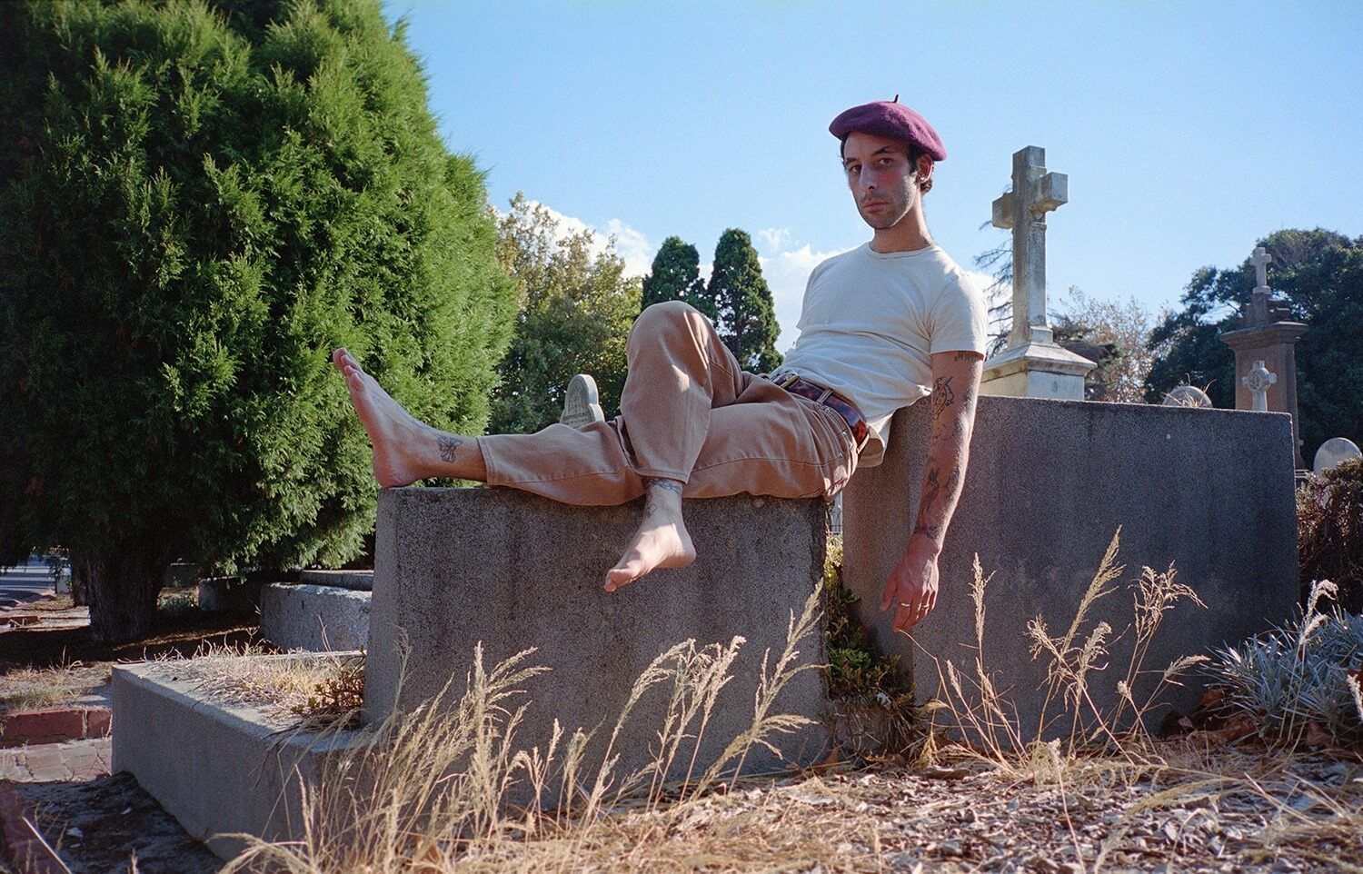 Gregor is wearing a red hat, sitting on a rock outdoors, crossing his legs, his sitting pose leans slightly backward.