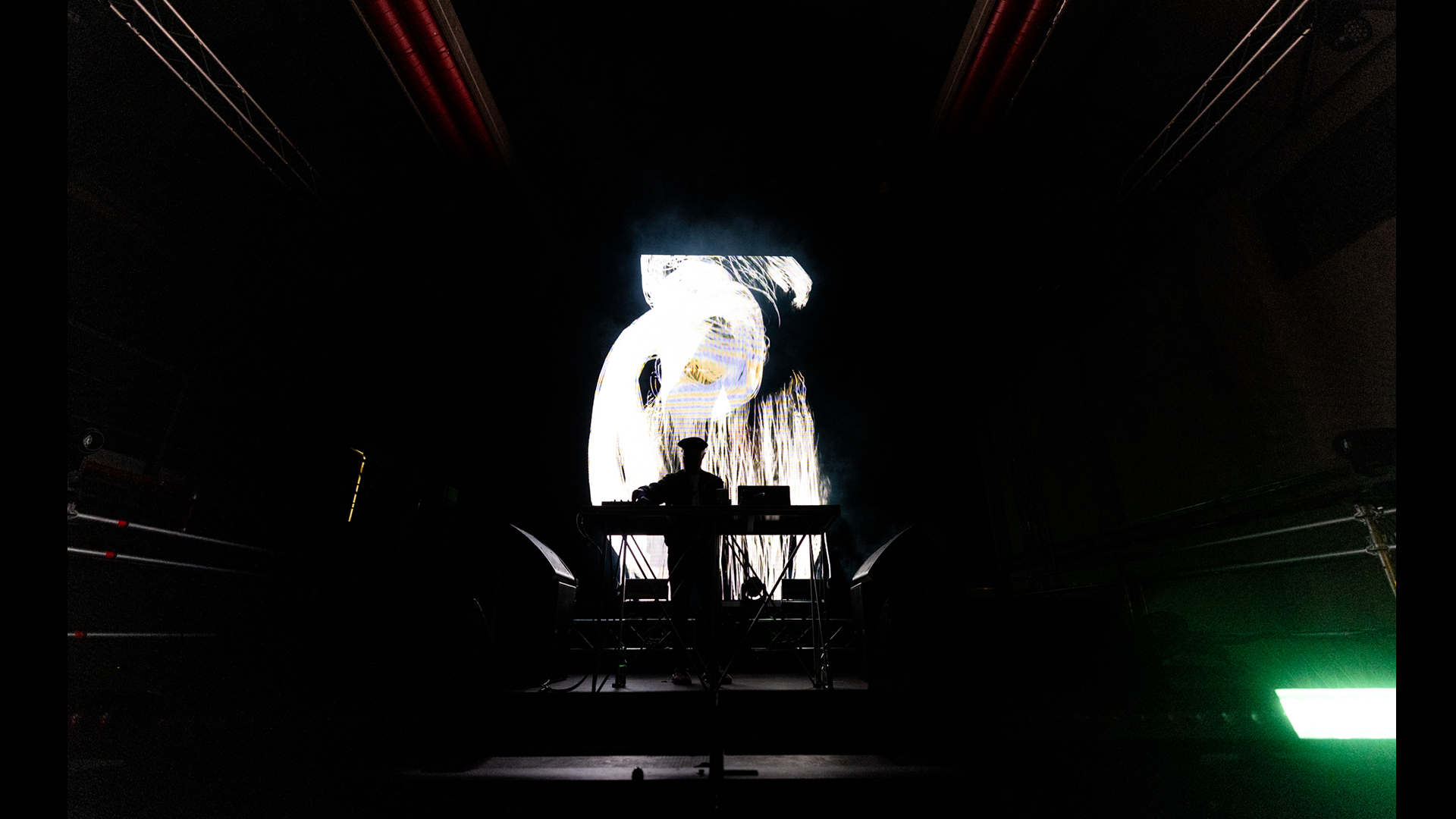 Forest Swords stand behind a DJ booth, silhouetted against a large abstract visual projection on stage.