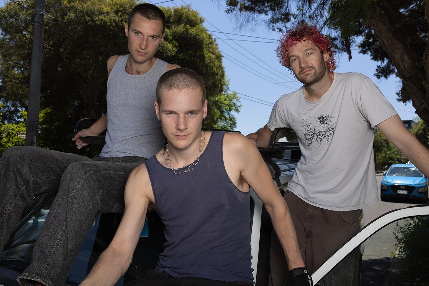 Three men posing together. The person in the front and on the left is wearing a tank top, with the person on the left sitting. The man on the right is wearing a T-shirt