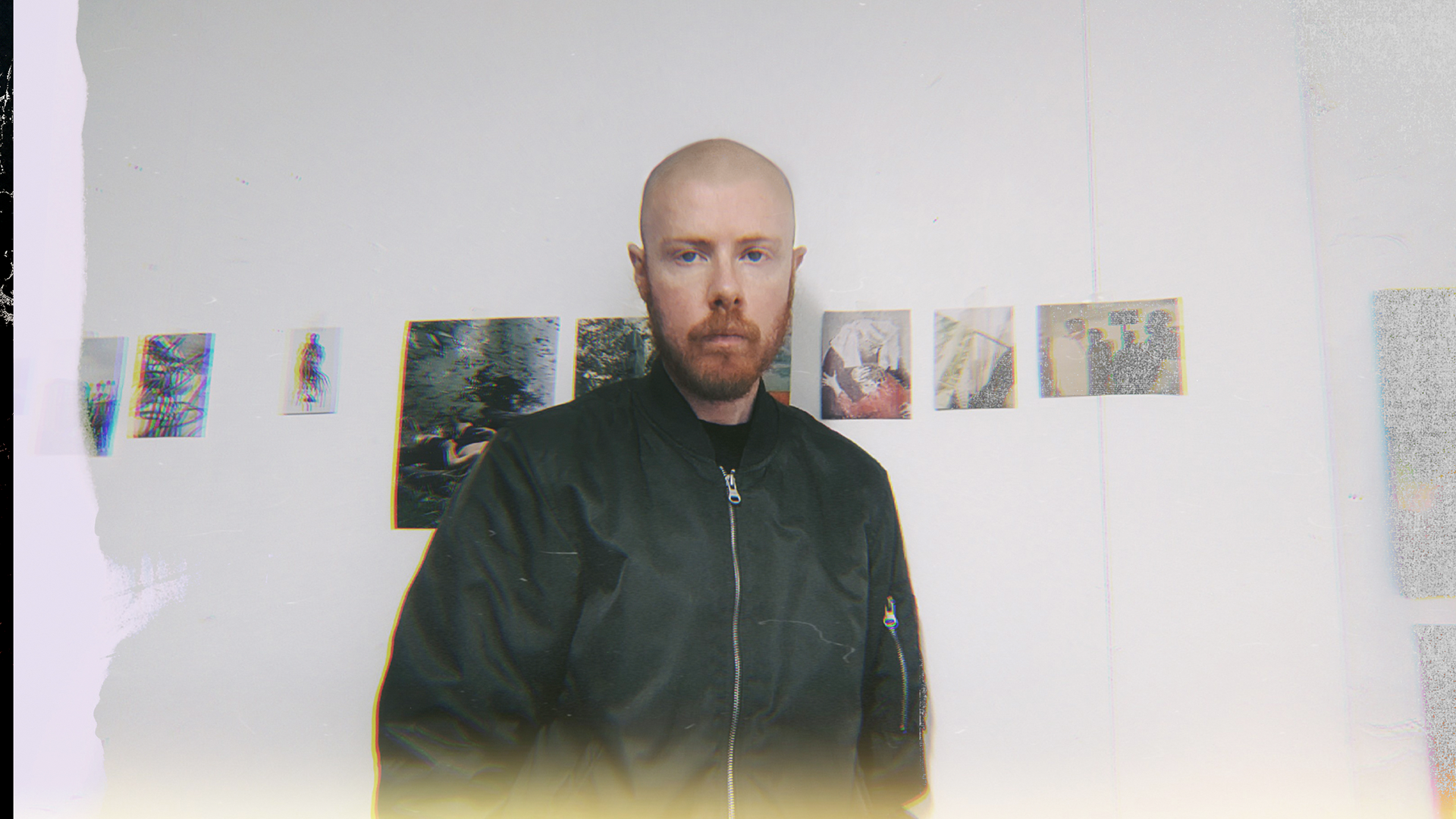 A person with shaved head and a beard stands in front of a white wall displaying small artworks.