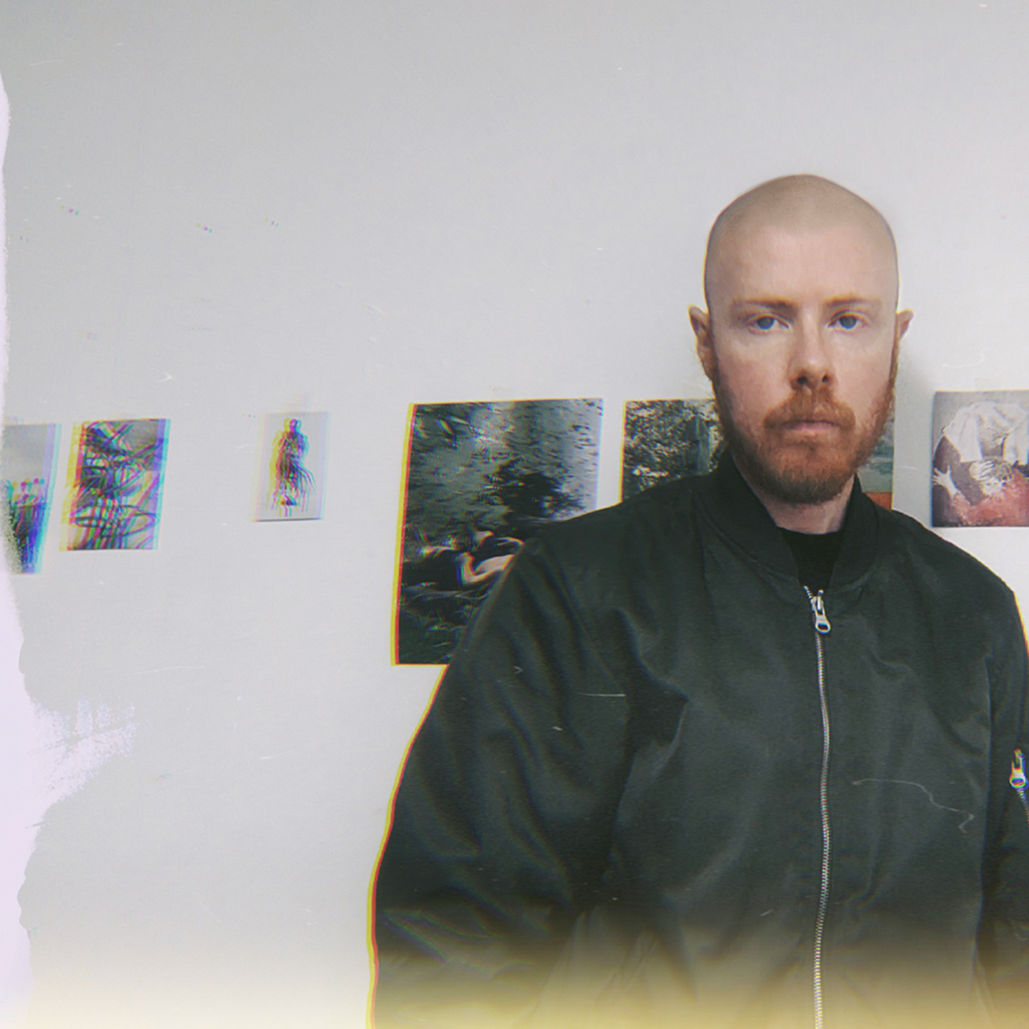 A person with shaved head and a beard stands in front of a white wall displaying small artworks.