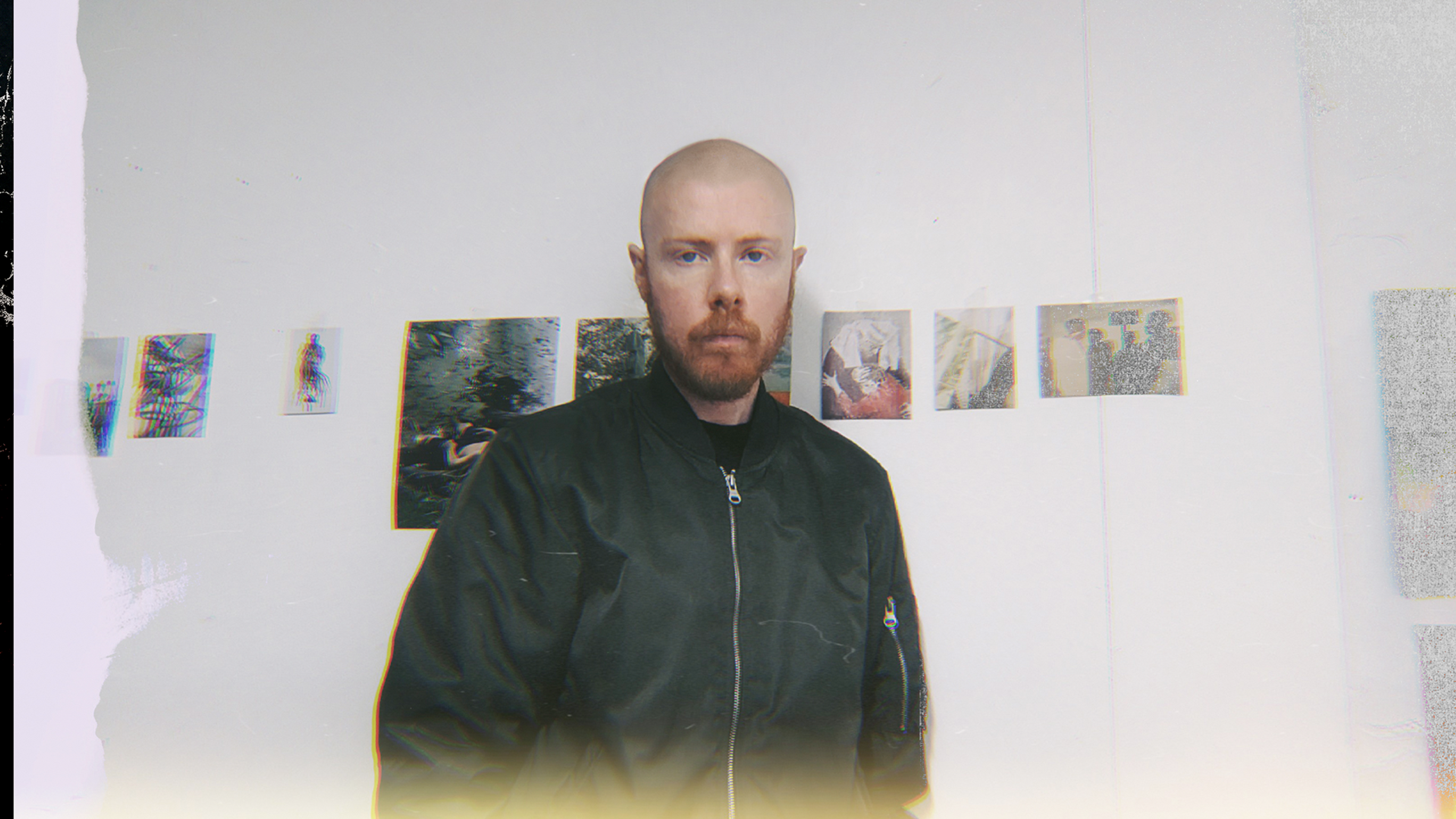 A person with shaved head and a beard stands in front of a white wall displaying small artworks.