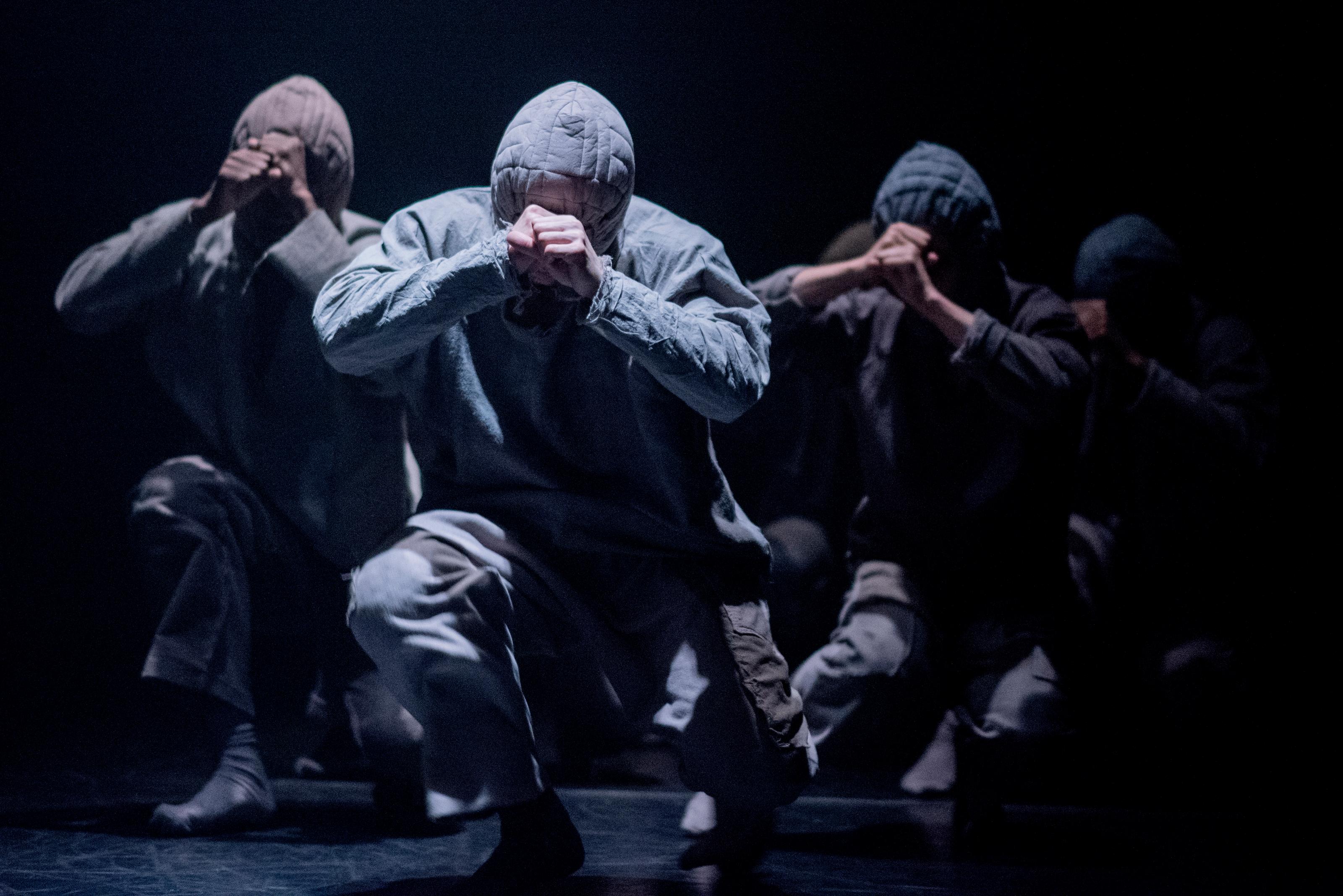 Four dancers in hoodies move towards the camera in a crouched position