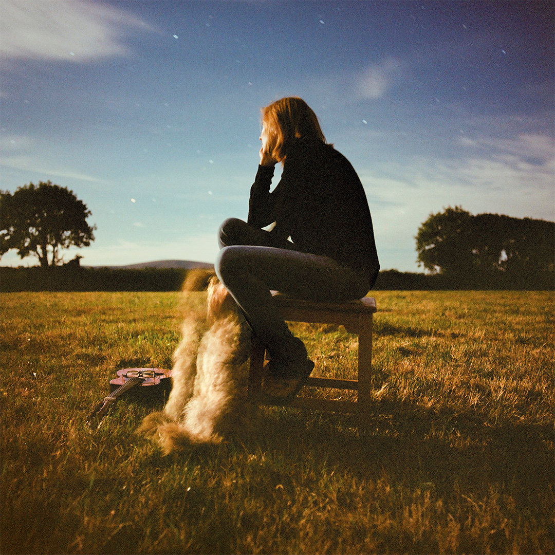 Woman sits on a stool, looking away from the camera towards a field. Her legs rest on the dog sitting in front of her.  