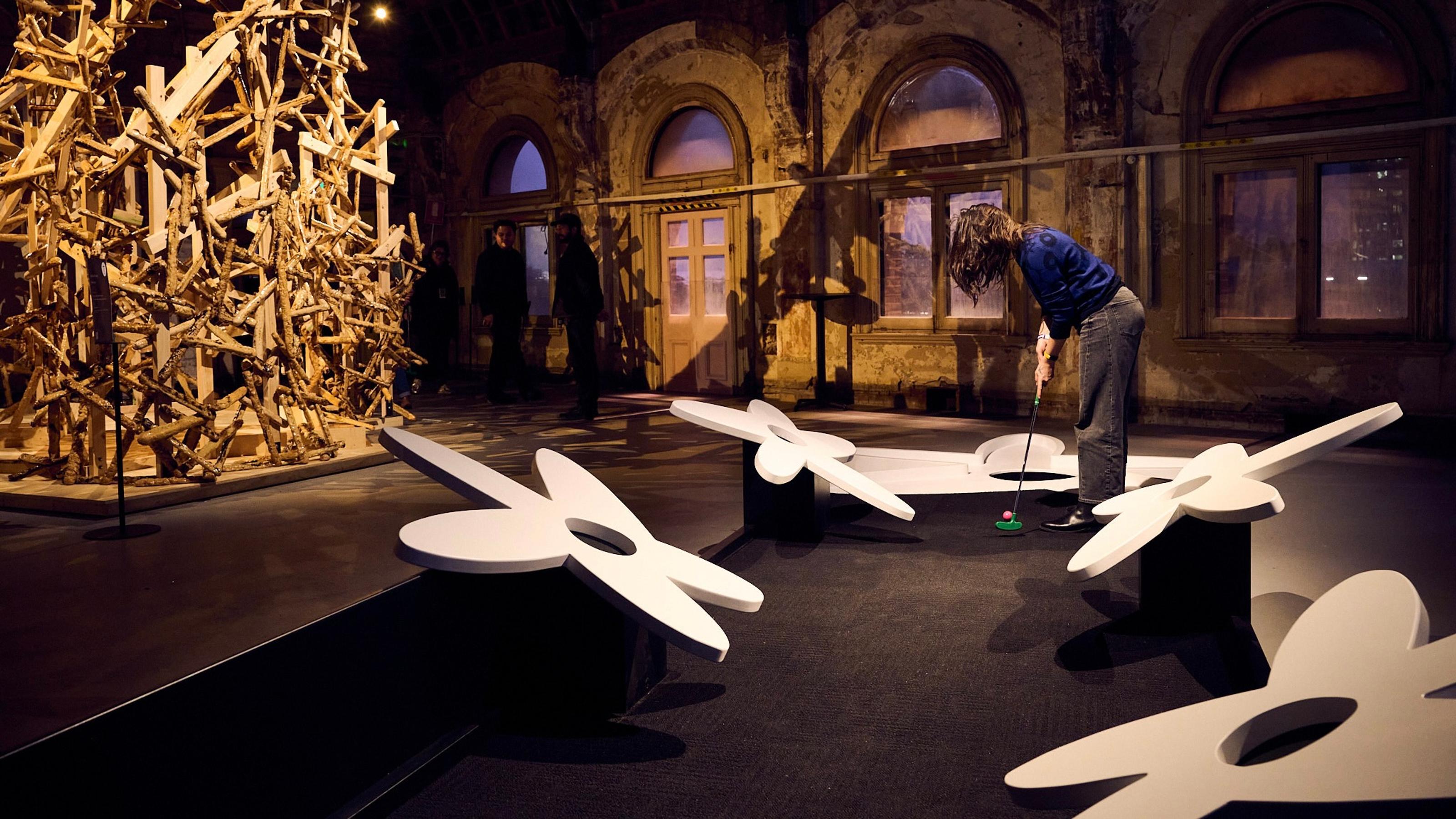Someone lines up to try the Swampflower mini golf hole, the carpet is flanked by cartoonish sculptures of flowers.