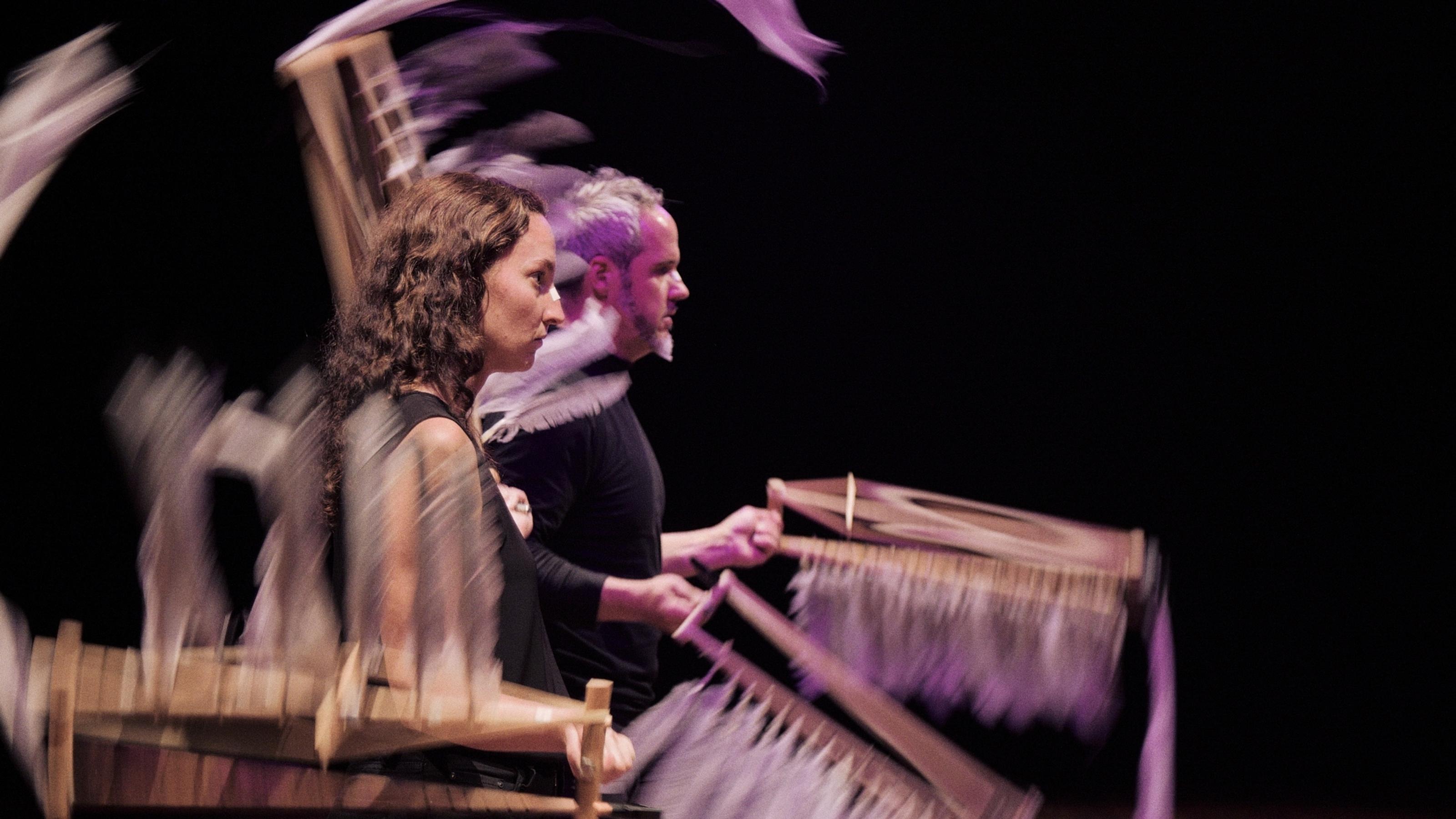 Two people stand on a dark stage and twirl feathered wings as they stare out to the audience.