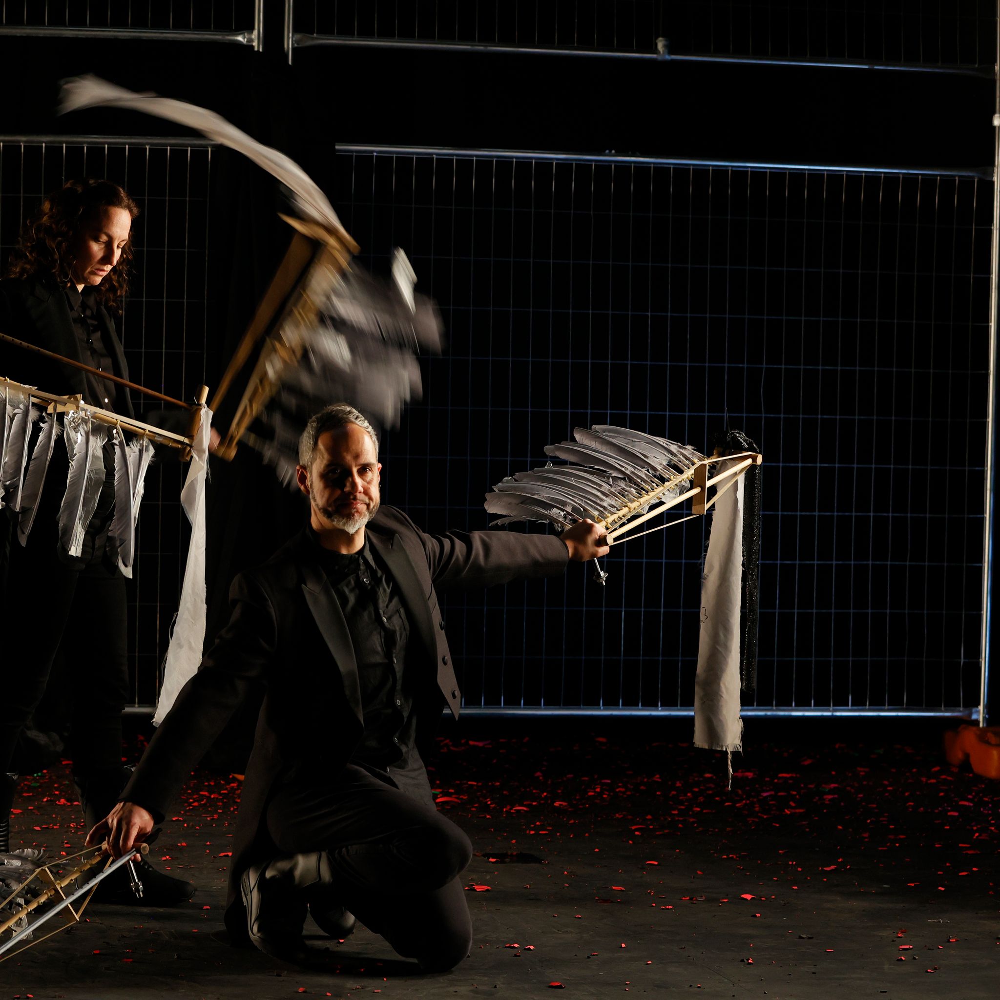 Two people, one standing and one kneeling in front of the other both wave feathered instruments around like wings.