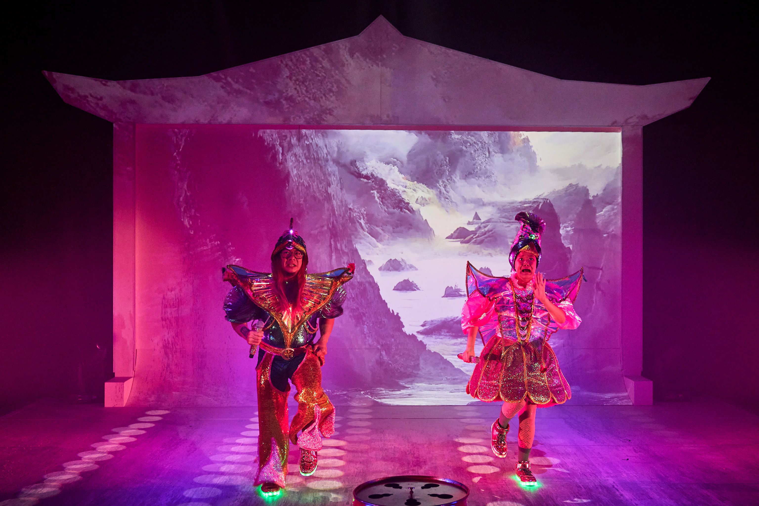 Joe Paradise Lui and Merlynn Tong look scared as they run, they are dressed in neon versions of ancient Chinese garments