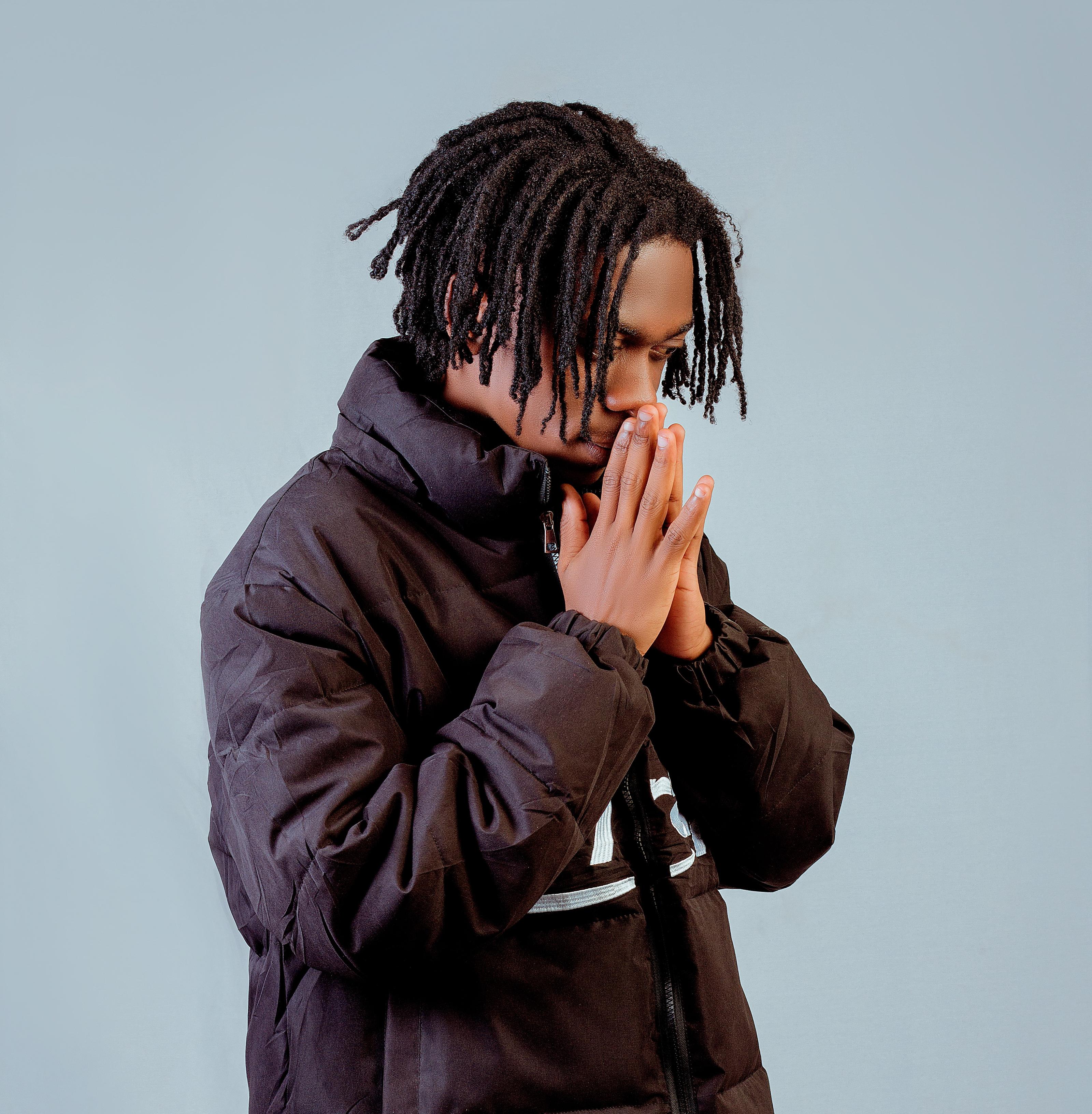 A person with short dreadlocks in a black jacket stands with hands pressed together against a plain background.