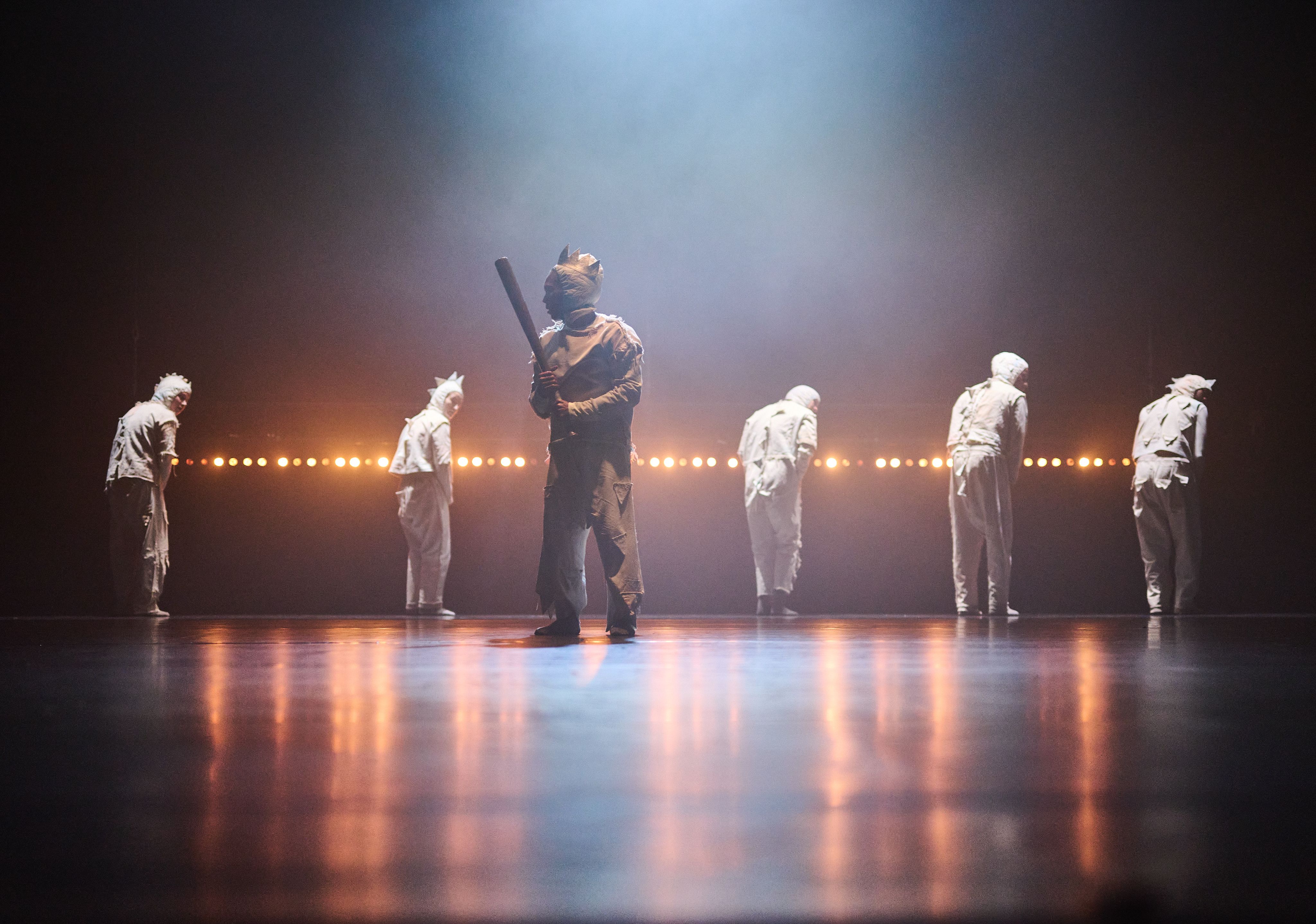 Five dancers in light tracksuits turn away from the camera in the background, in the foreground. Another dancer holding a baseball bat stands in the foreground 