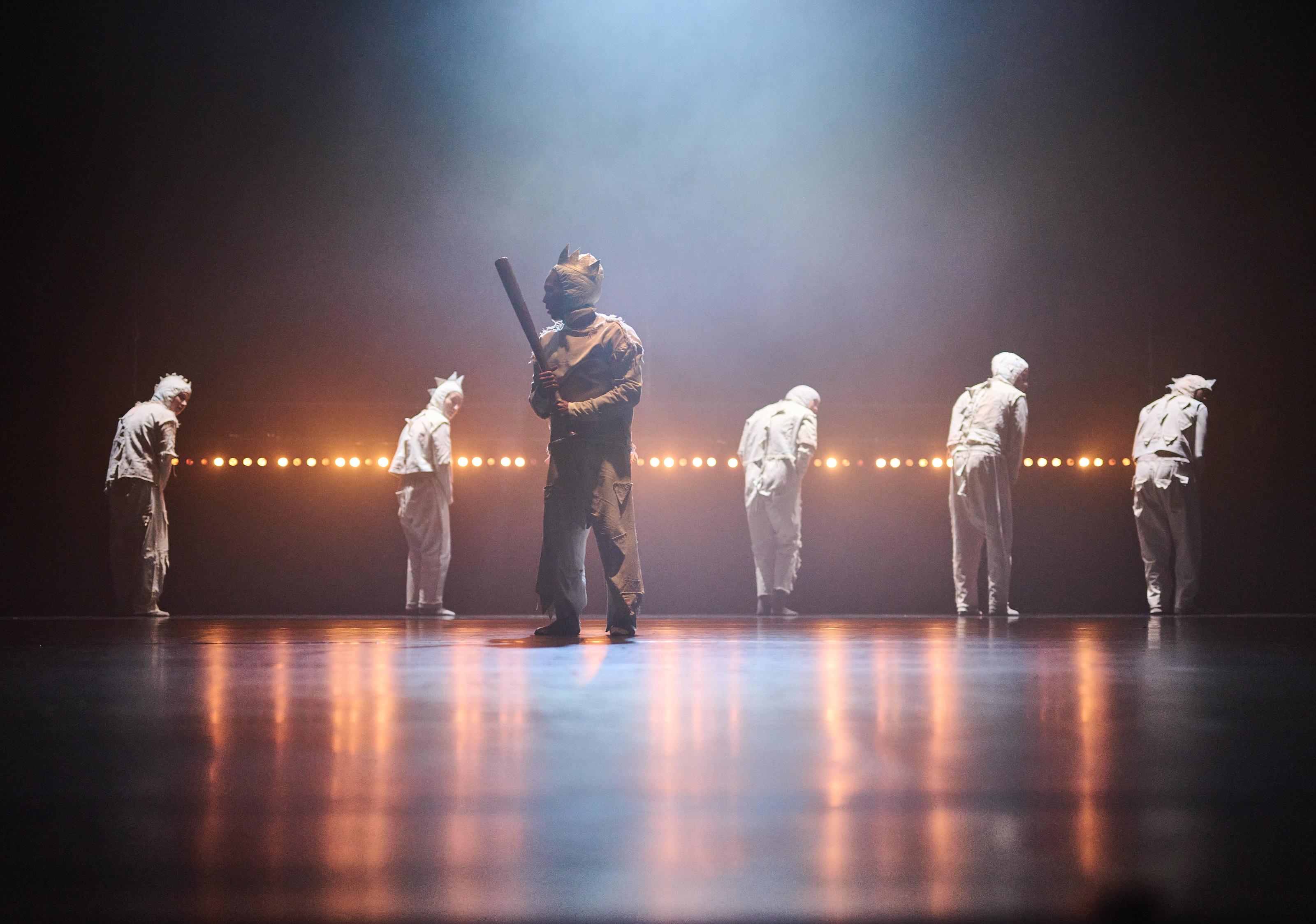 Five dancers in light tracksuits turn away from the camera in the background, in the foreground. Another dancer holding a baseball bat stands in the foreground