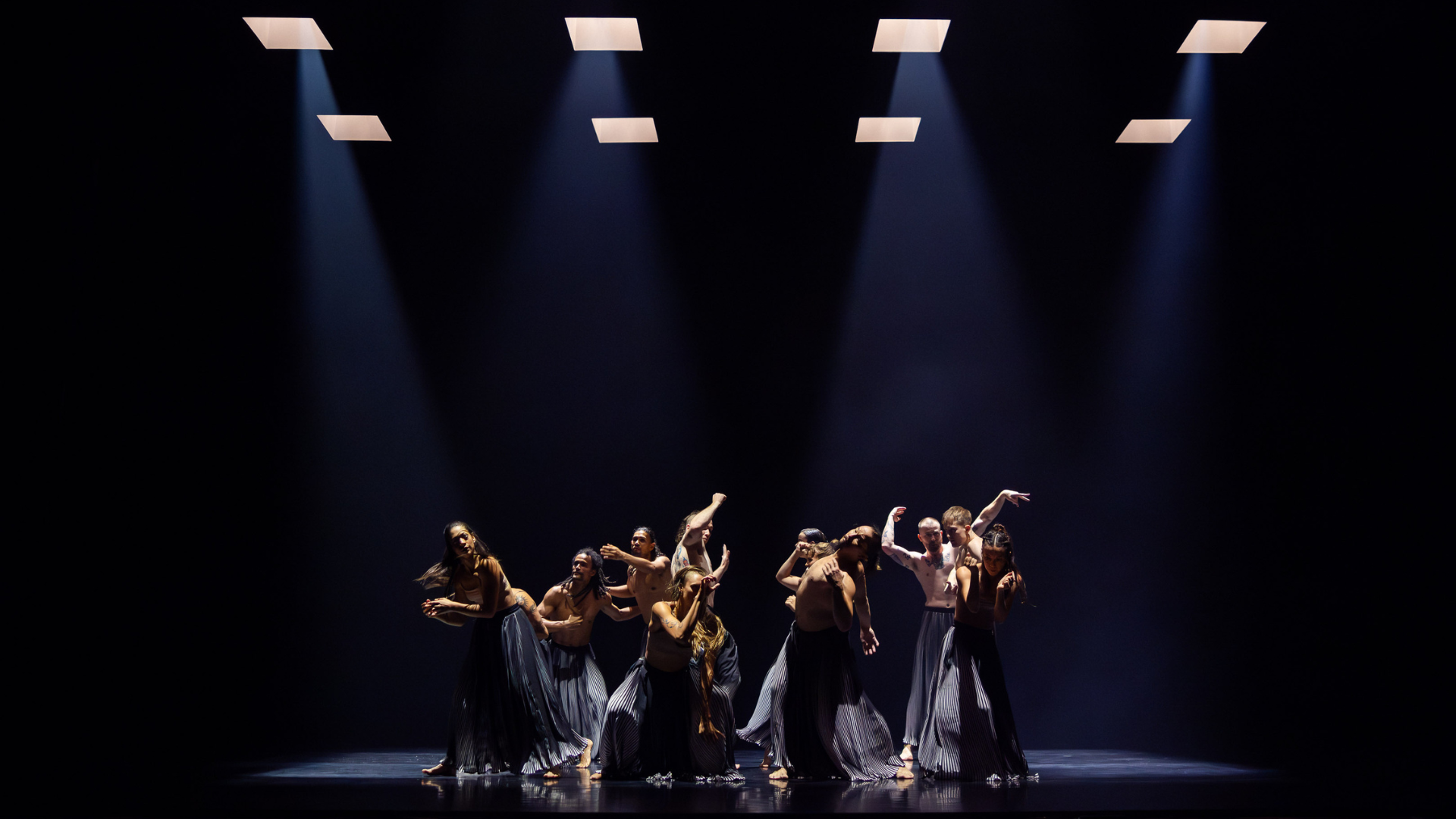 Many people dance in the centre of a stage, each in unique positions, illuminated by the four spotlights above them.