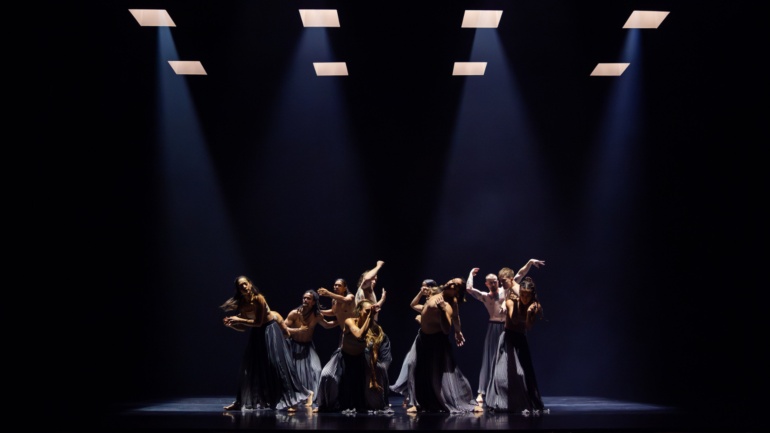 Many people dance in the centre of a stage, each in unique positions, illuminated by the four spotlights above them.