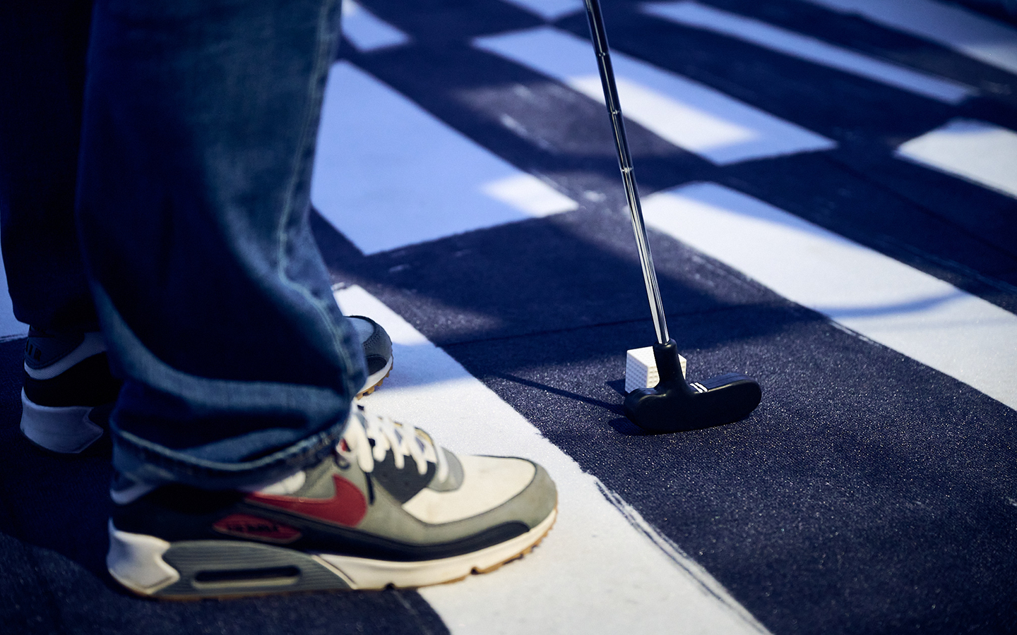 A close-up view of feet and a mini golf putter, putting a square ball, in Delaine Le Bas's installation 'Square peg, round hole. NO!'